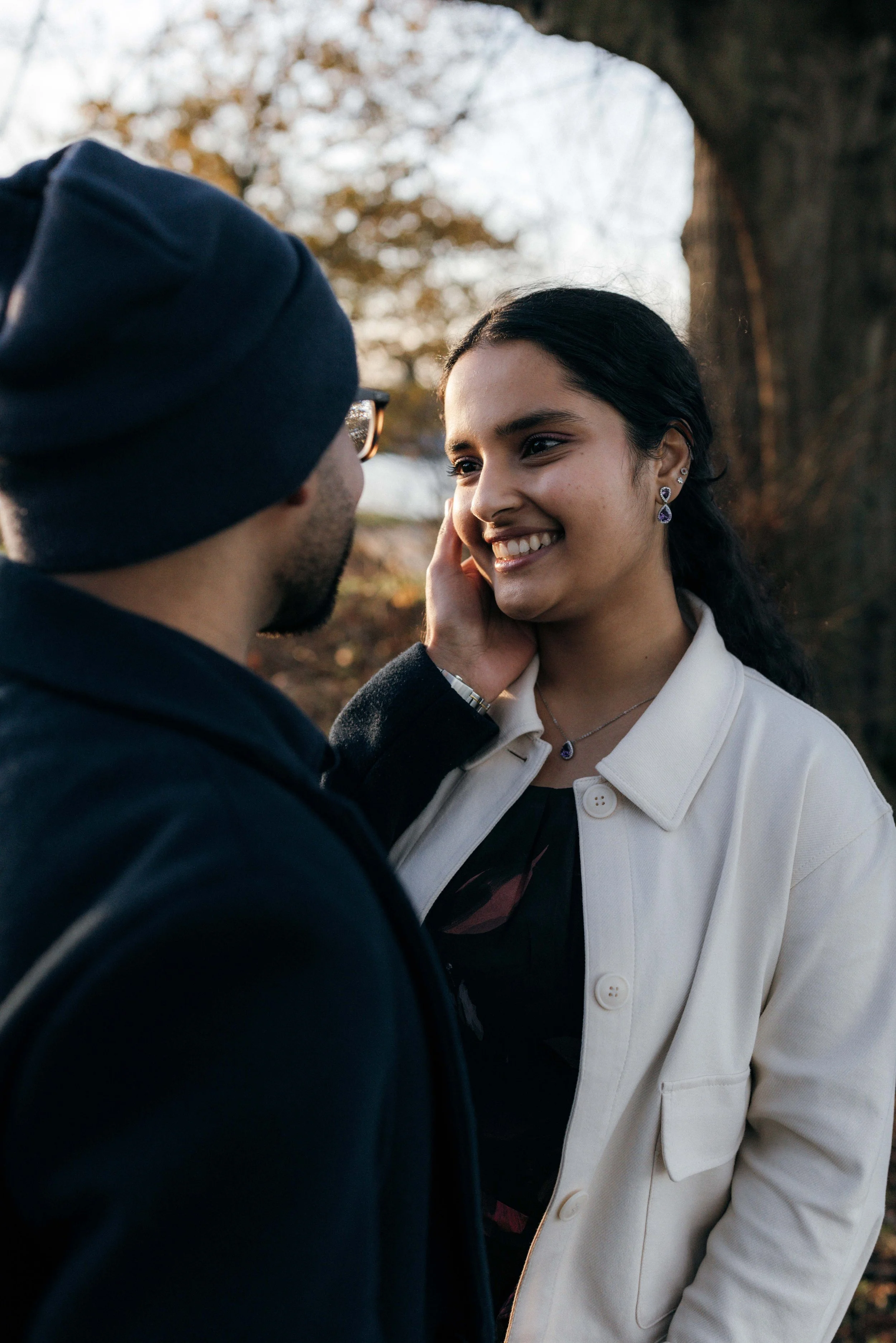 A man and woman sharing an intimate moment outdoors, with the man gently touching the woman's face as she smiles warmly at him. The background features trees and a clear sky.
