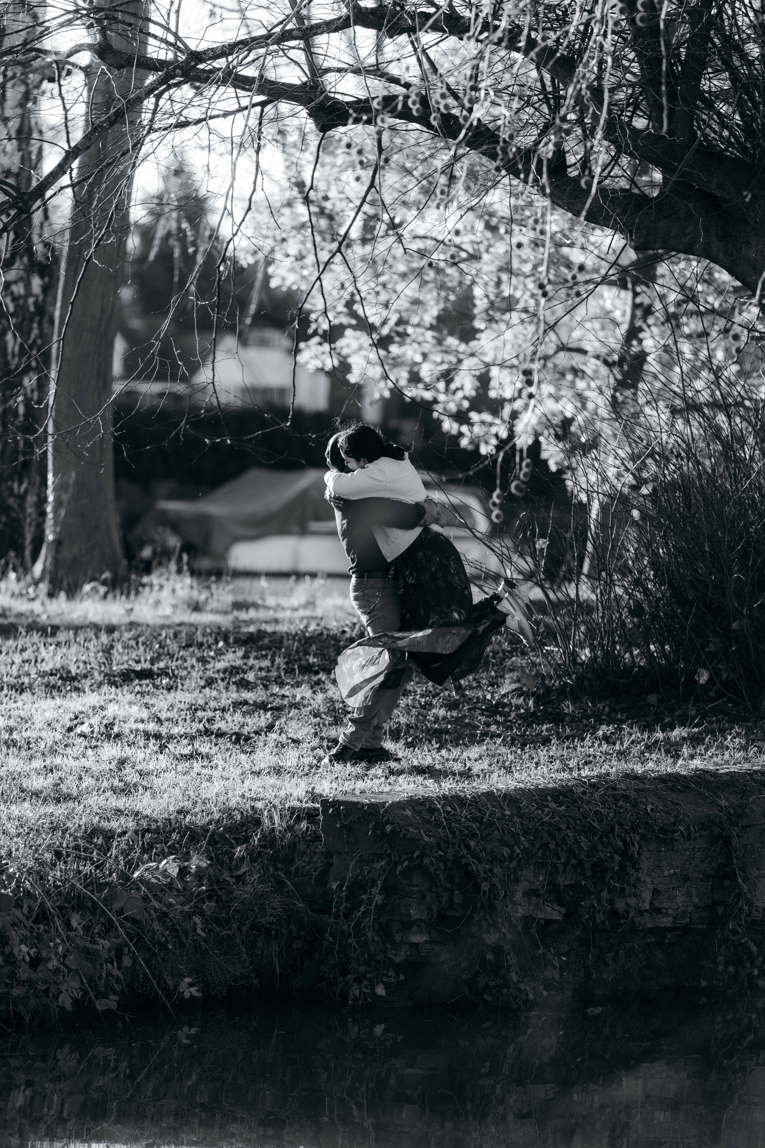 A couple embraces near a tree, with one person lifting the other, set outdoors near a body of water, with sunlight filtering through the branches.