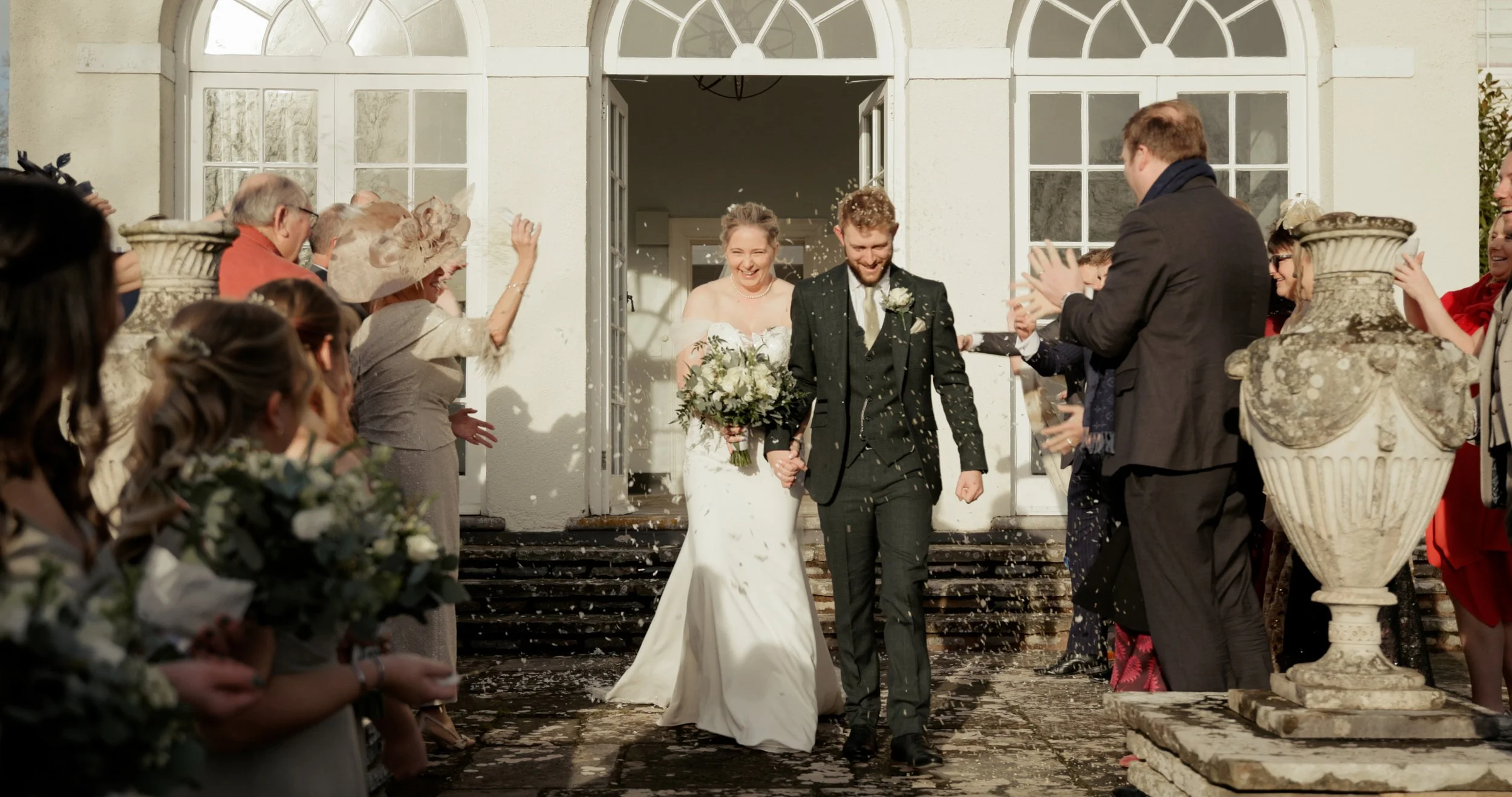 A newlywed couple walking out of a building, surrounded by cheering friends and family, celebrating with confetti, on the steps of a white building with large windows.