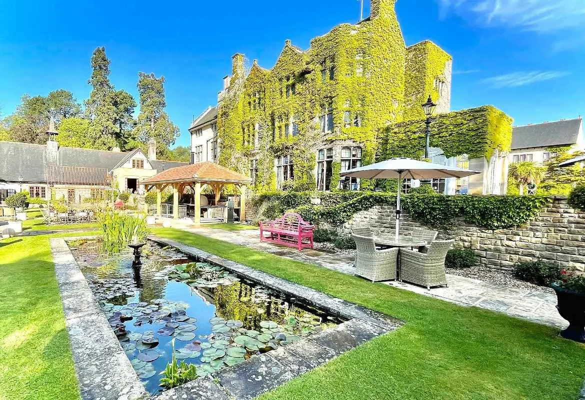 Beautiful garden with a pond, patio furniture, and a historic ivy-covered castle in the background on a sunny day.