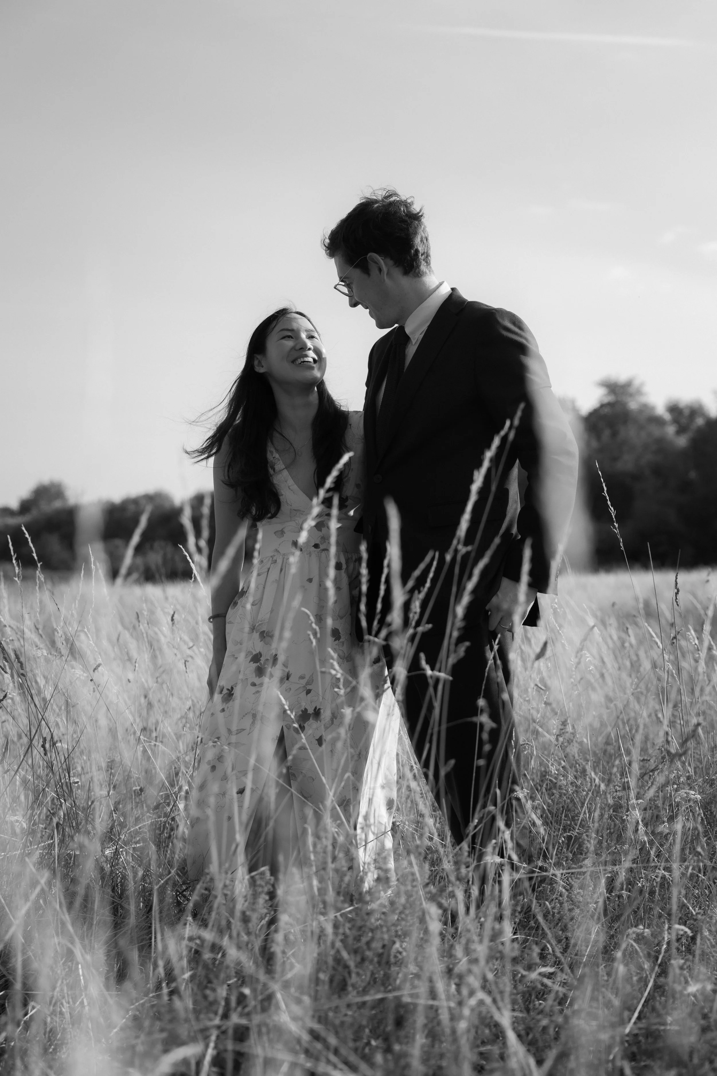 A black-and-white photo of a couple standing in a grassy field, smiling and looking at each other.