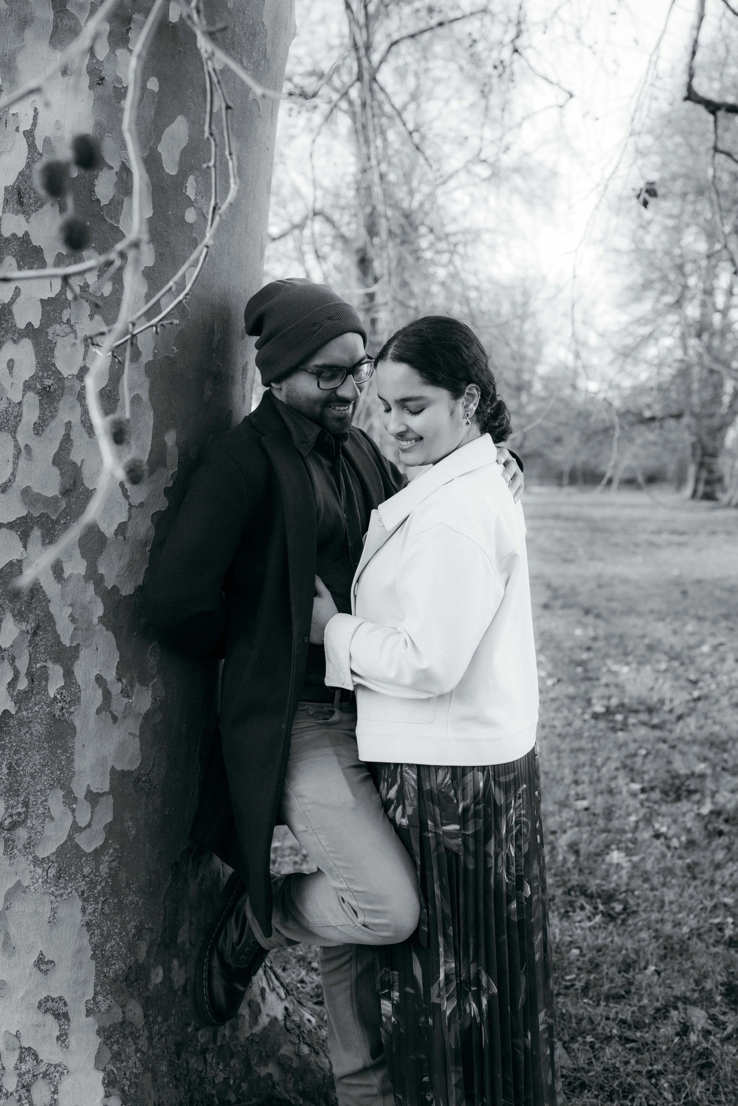 A couple smiling and leaning against a tree outdoors in a park, with autumn trees in the background.