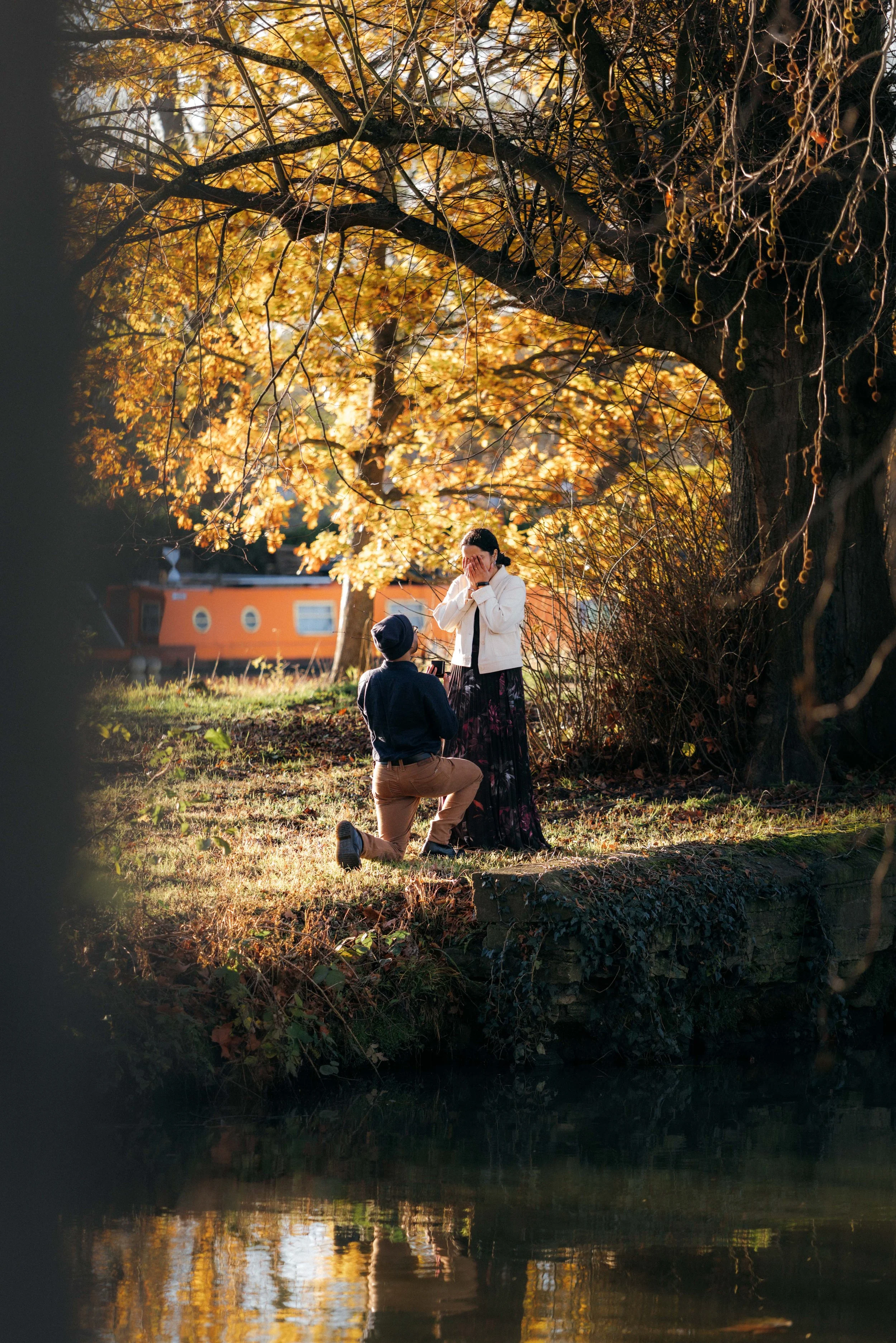 A man proposing marriage to a woman outdoors by a pond surrounded by autumn trees while a photographer captures the moment.