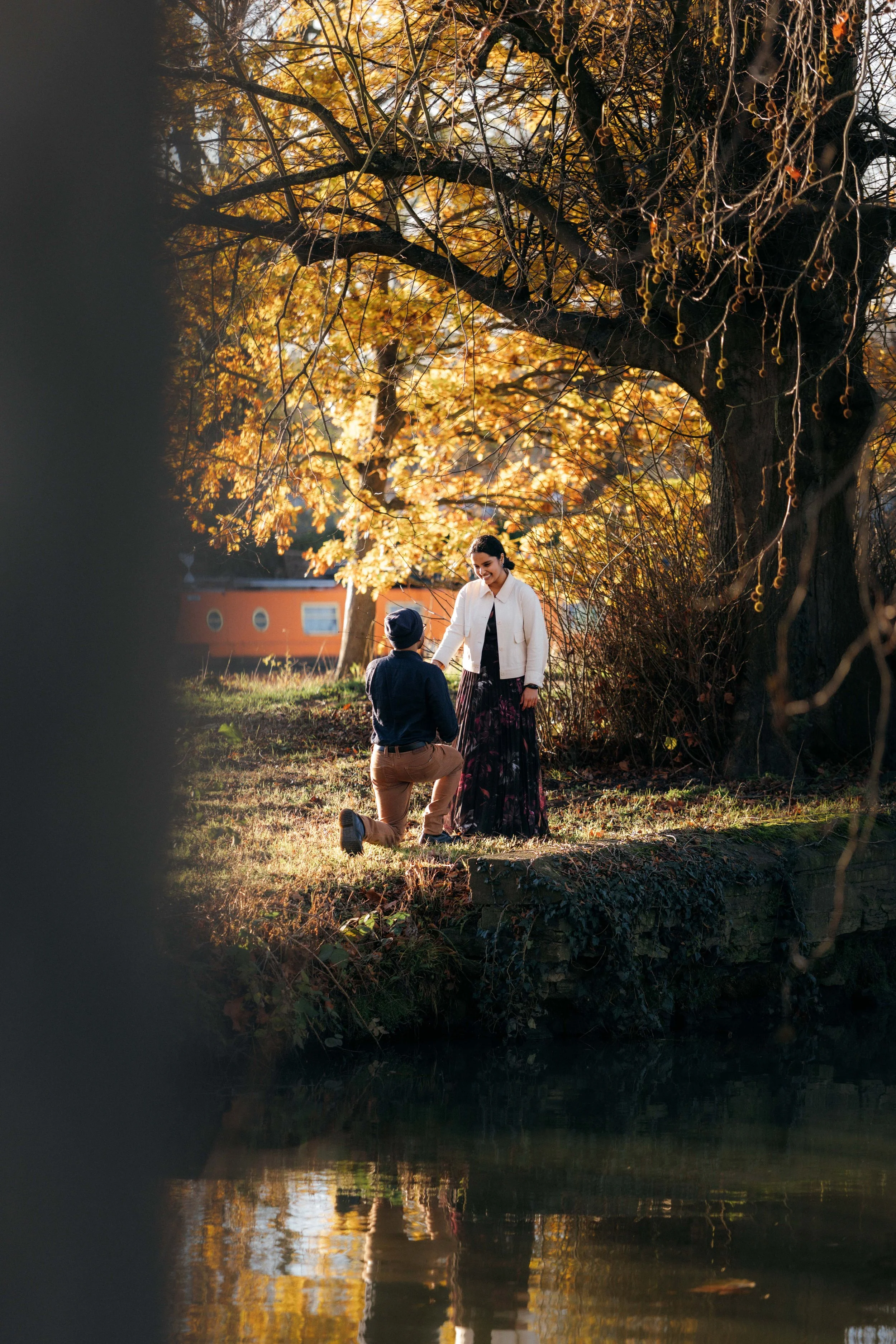 A man proposes to a woman by a river during autumn, with trees having yellow leaves and a small orange house in the background.