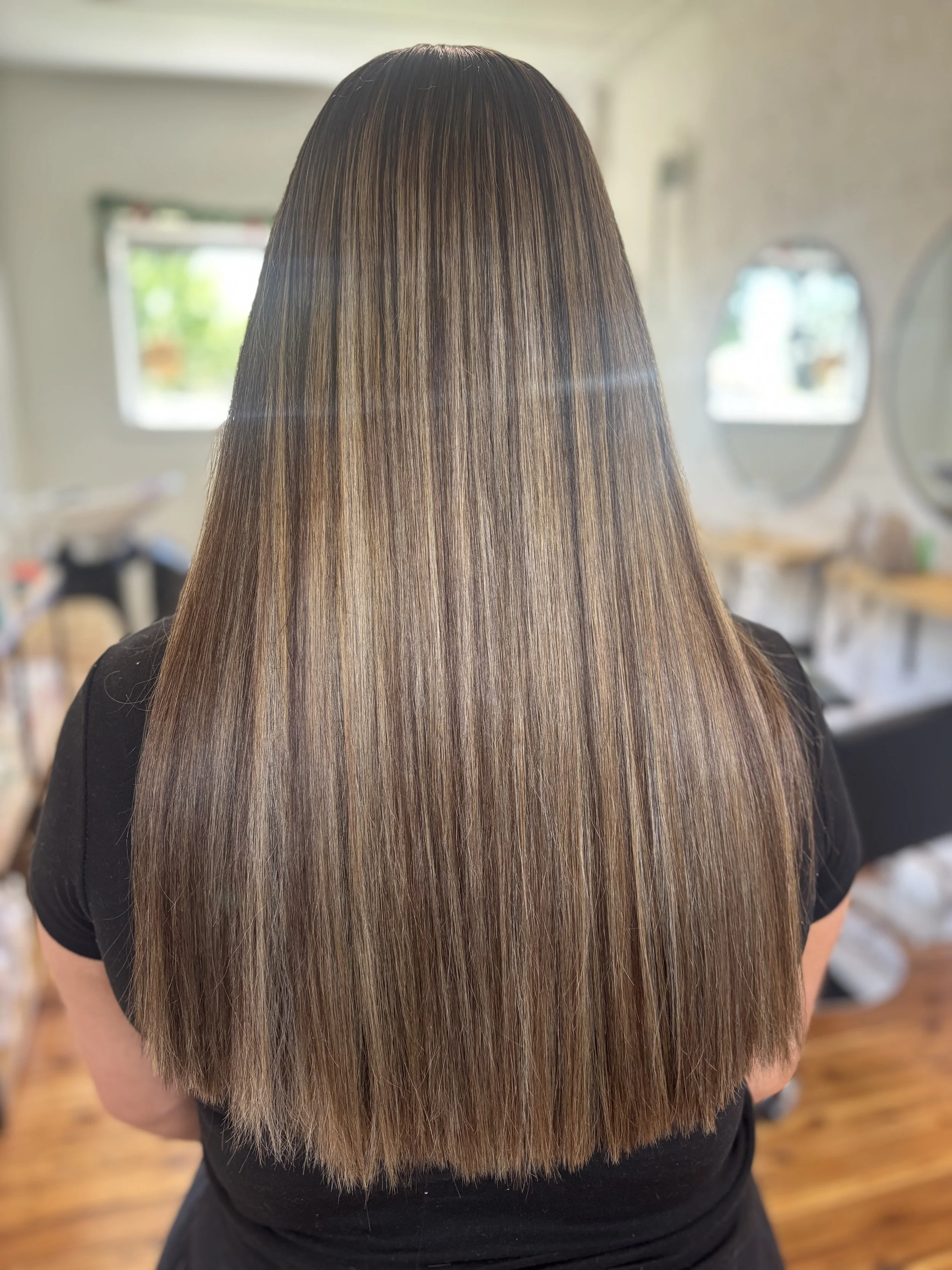 Back of a woman with long, straight, brown hair with subtle highlights, in a salon or a room with natural light and mirrors.