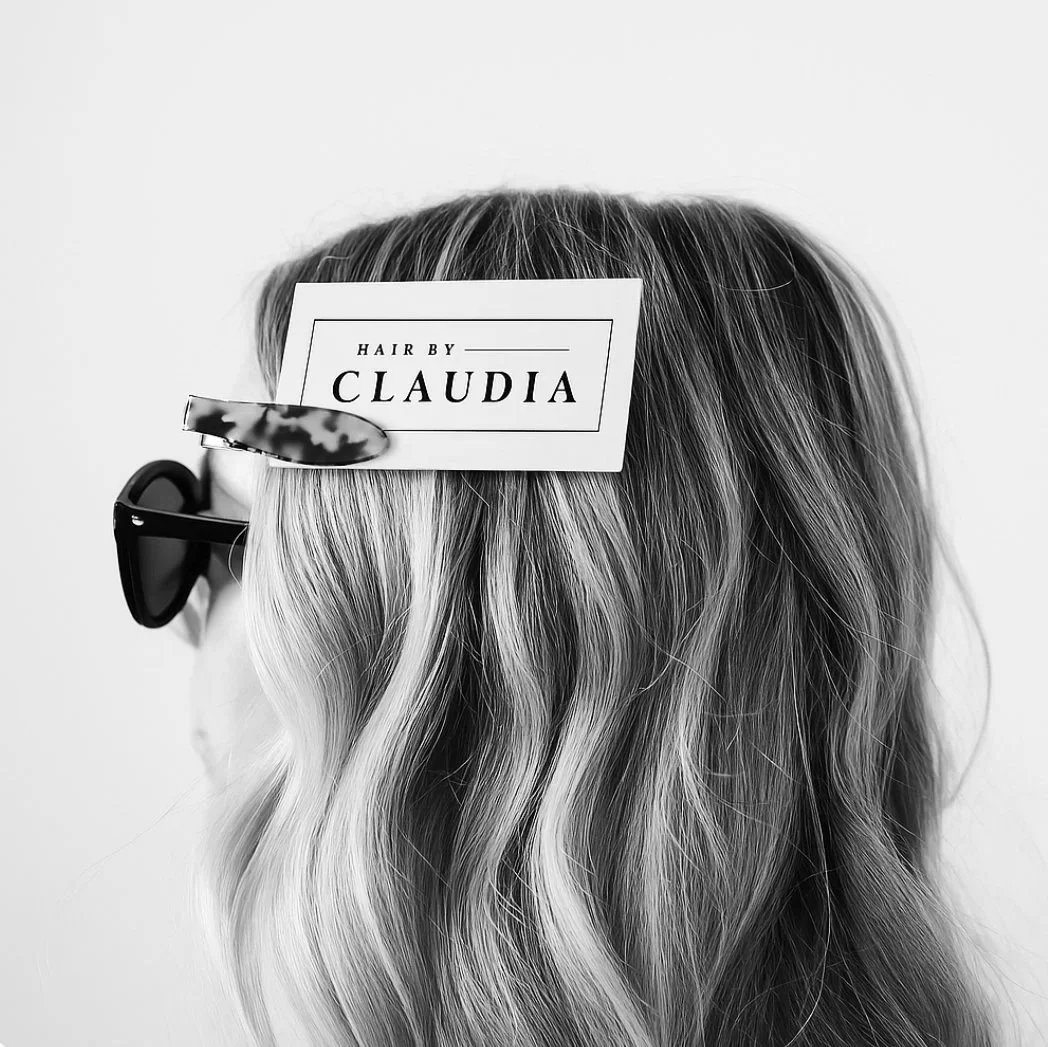 Black and white photo of a woman's head with wavy hair, wearing sunglasses. A label on her head reads 'Hair by Claudia'.