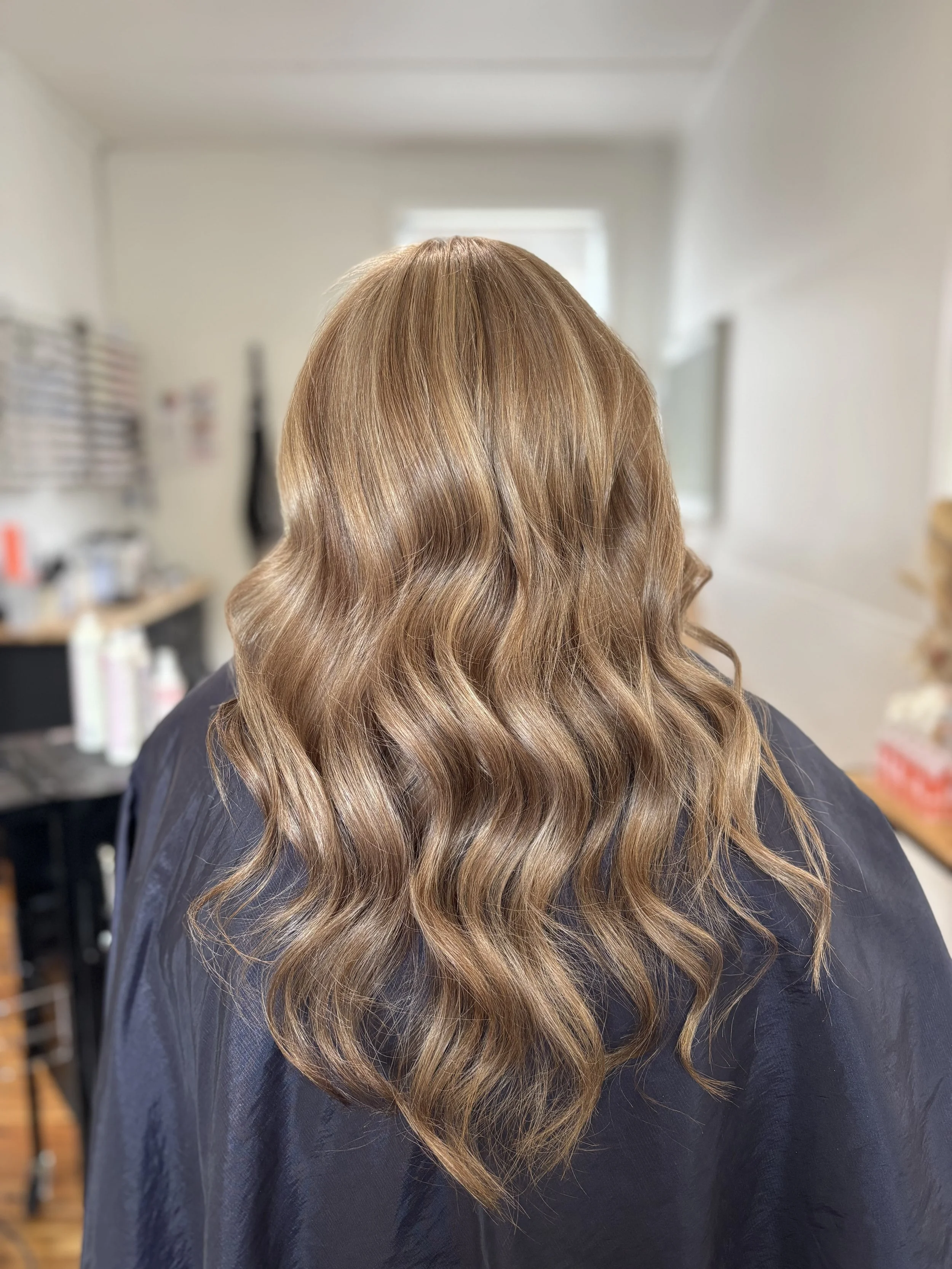 Back view of a person with long, wavy, light brown hair inside a salon, with a blurred background including salon tools and equipment.