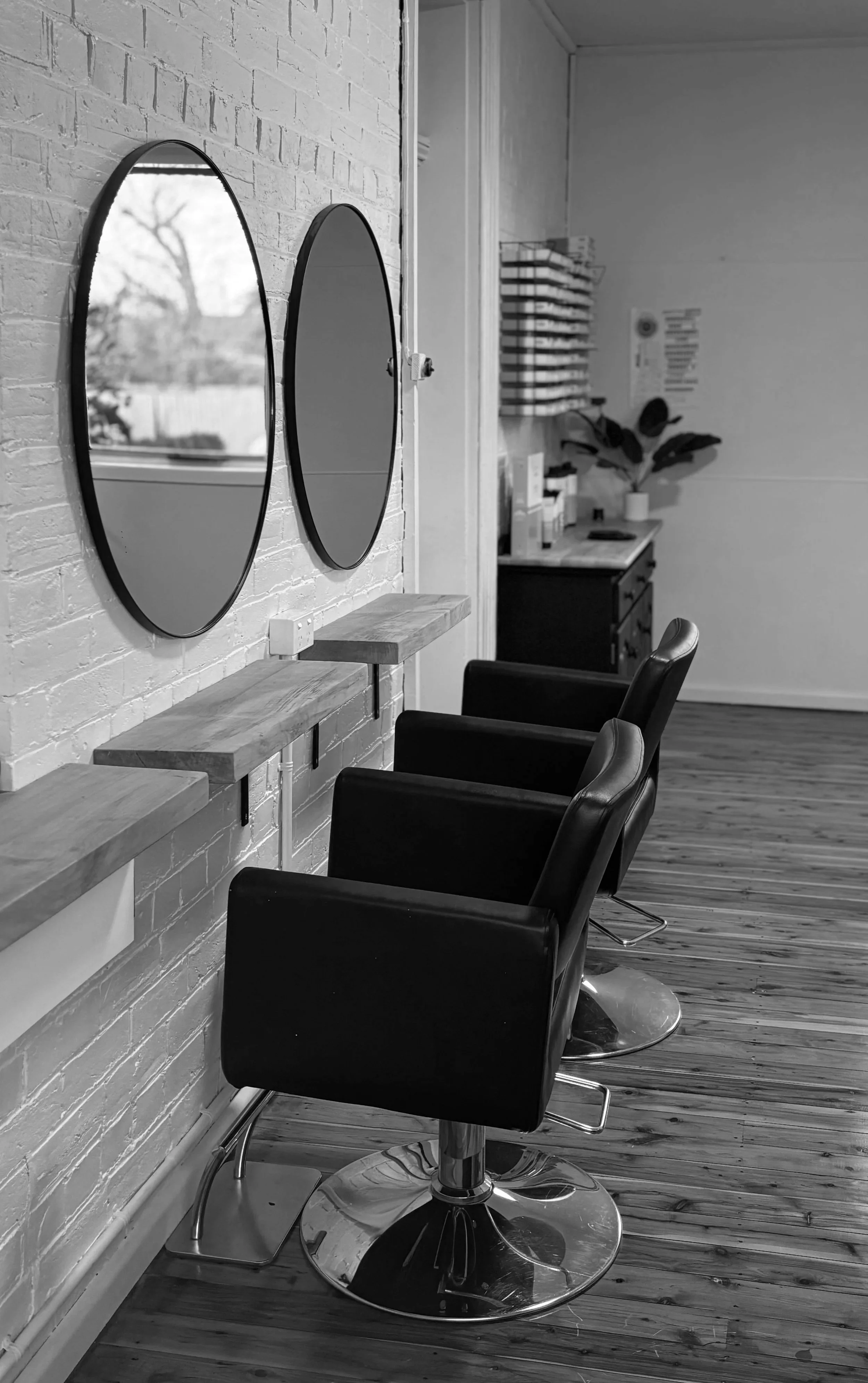 Black hair styling chairs in a salon with mirrors on a white brick wall, wooden shelves, and salon supplies in the background.