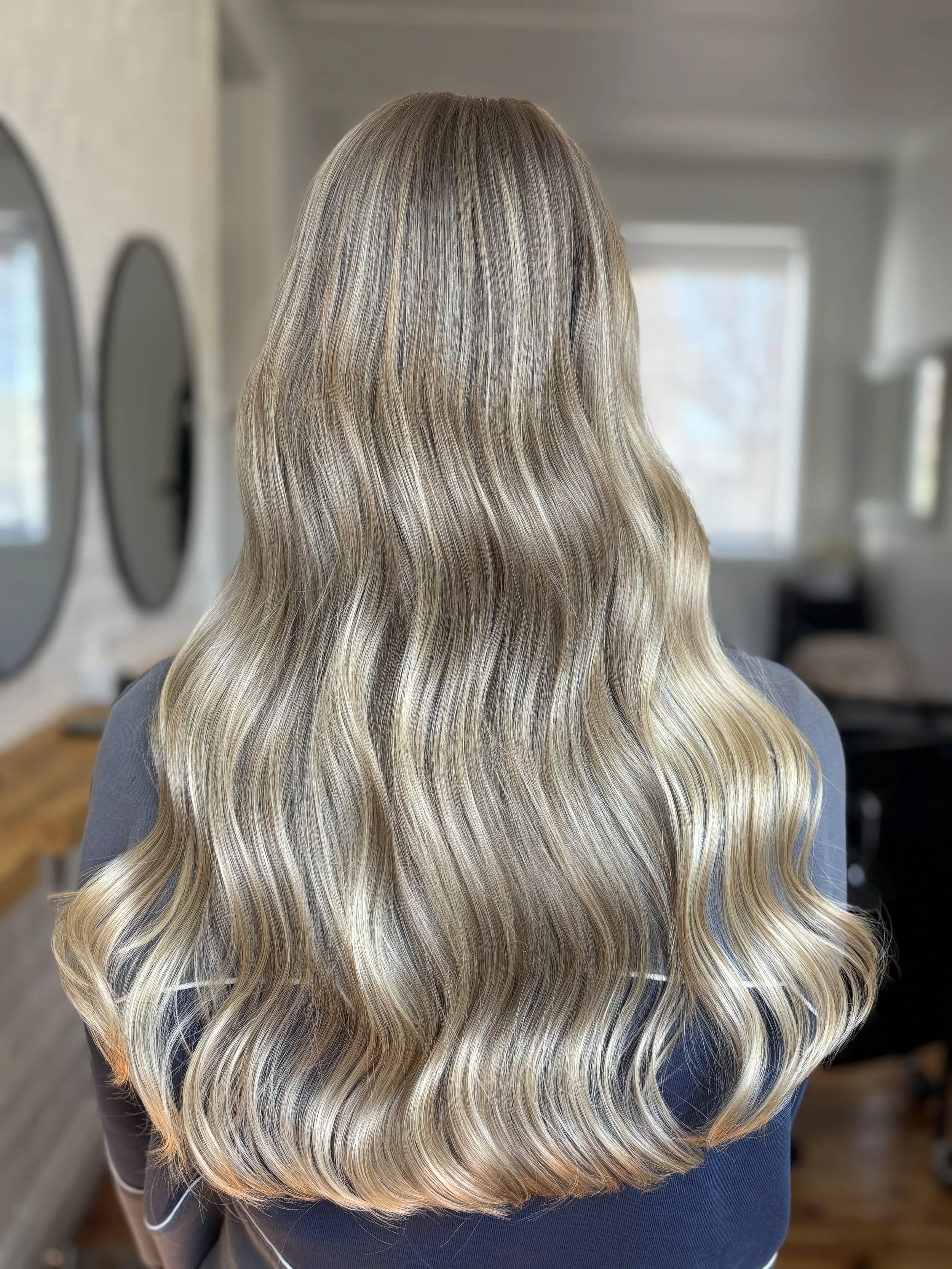 Back of a woman with long, wavy, blonde hair with highlights, indoors with mirrors and windows in the background.