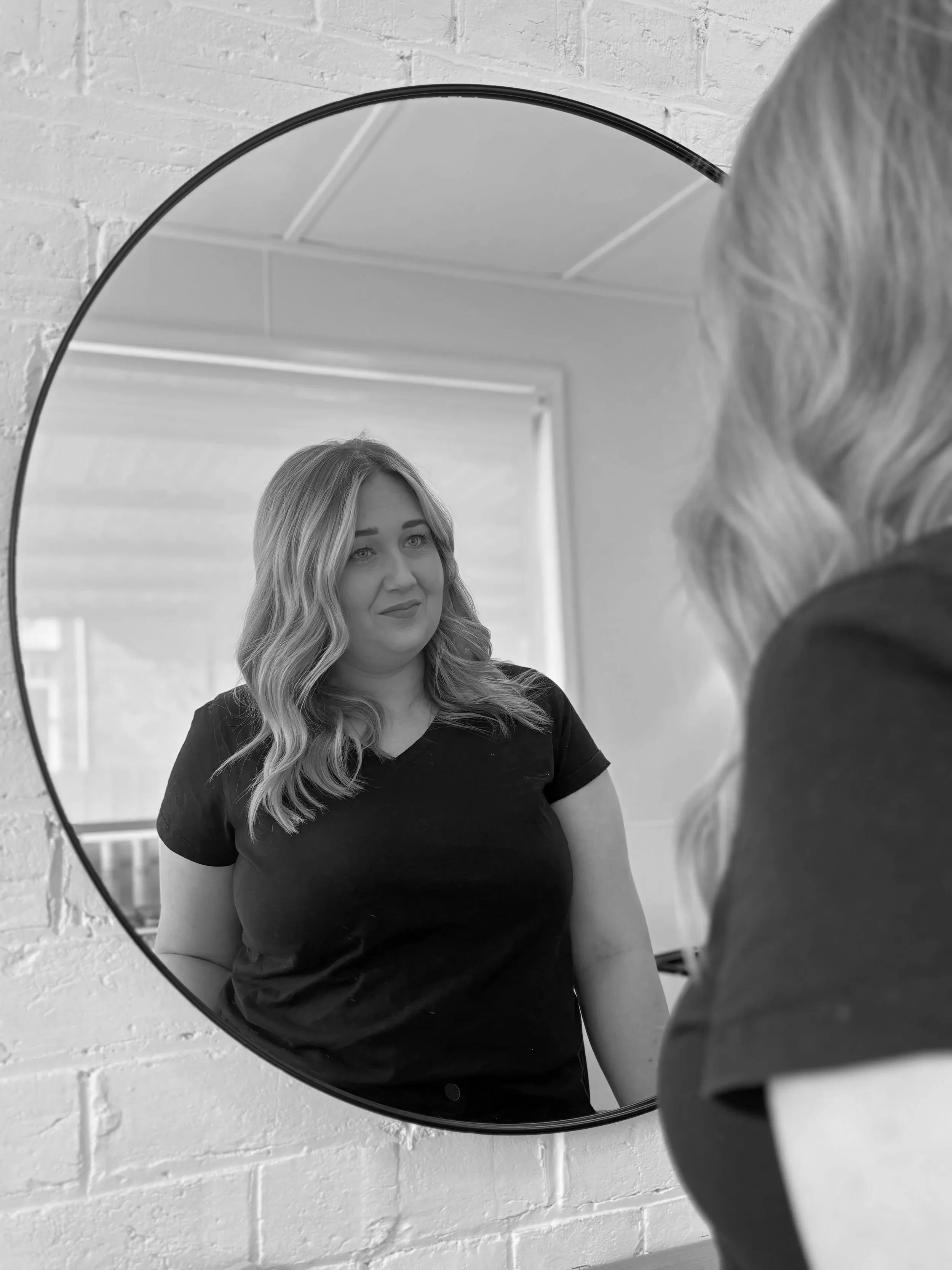 A woman with wavy blonde hair wearing a black t-shirt looks at herself in a circular mirror mounted on a white brick wall.