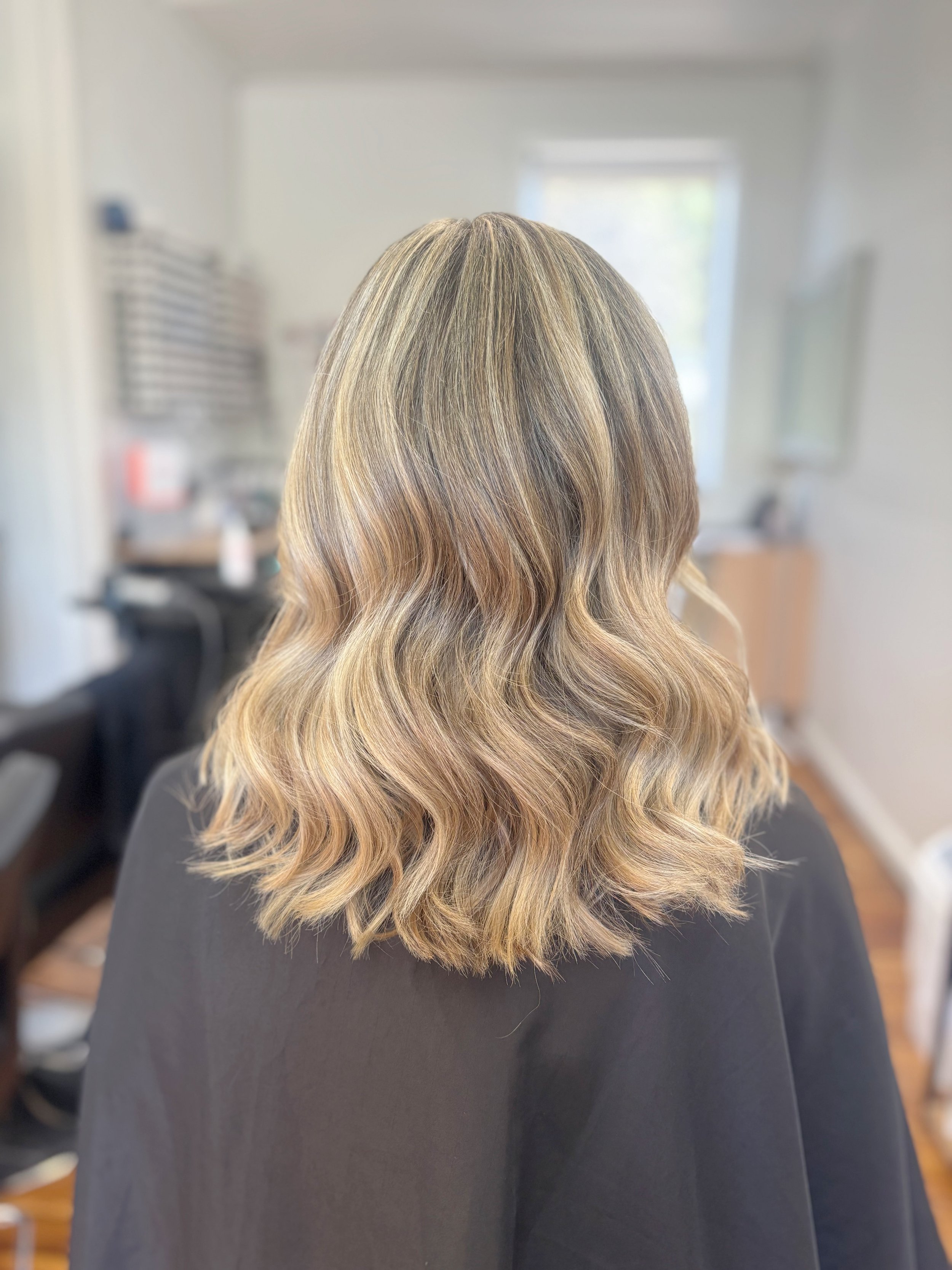 Woman with wavy, blonde hair seen from the back inside a salon or a room with a window, some furniture, and salon equipment.