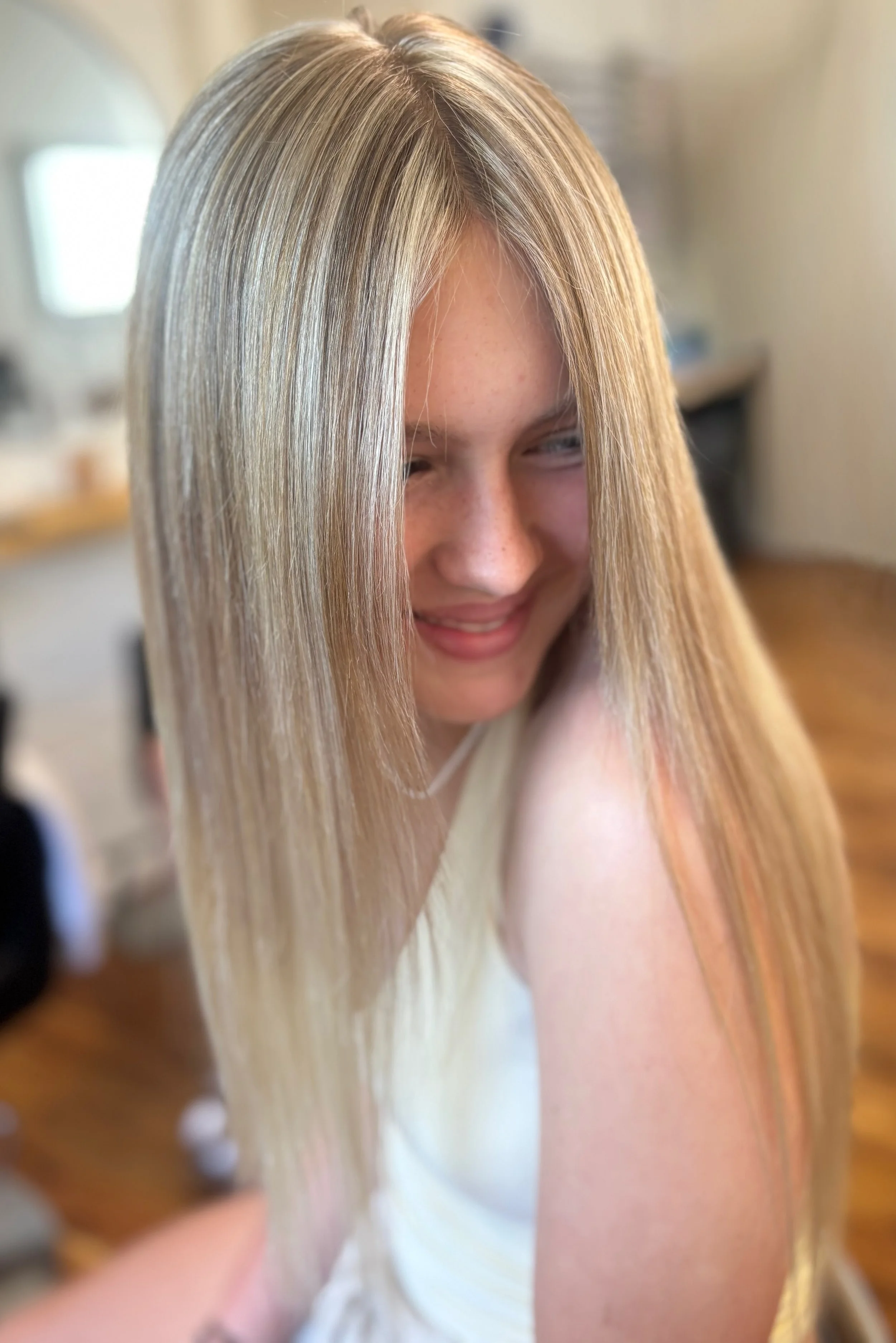 Close-up of a young woman with long, straight blonde hair smiling and looking down in a well-lit indoor setting.