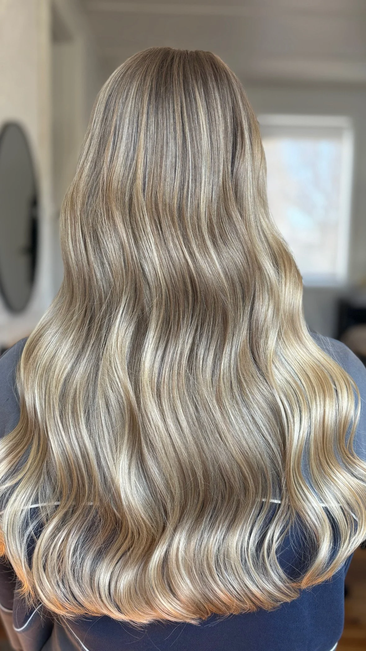 Back of a person with long, wavy blonde hair with highlights, in a well-lit room with a window in the background.