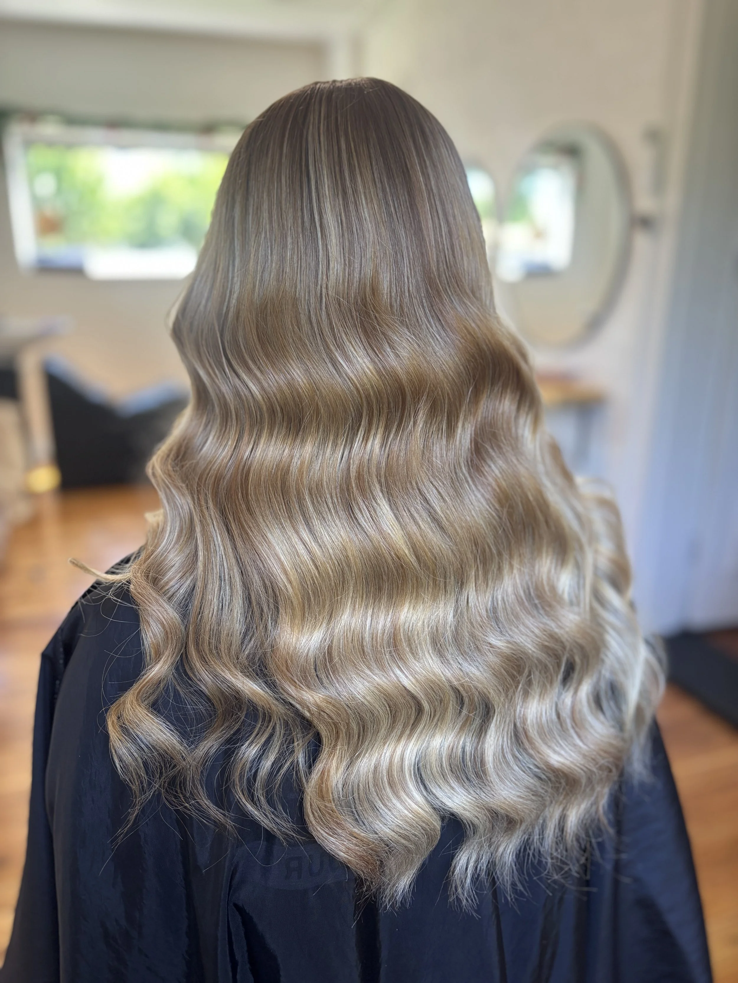 Back view of a person with long, wavy, blonde hair in a salon setting.