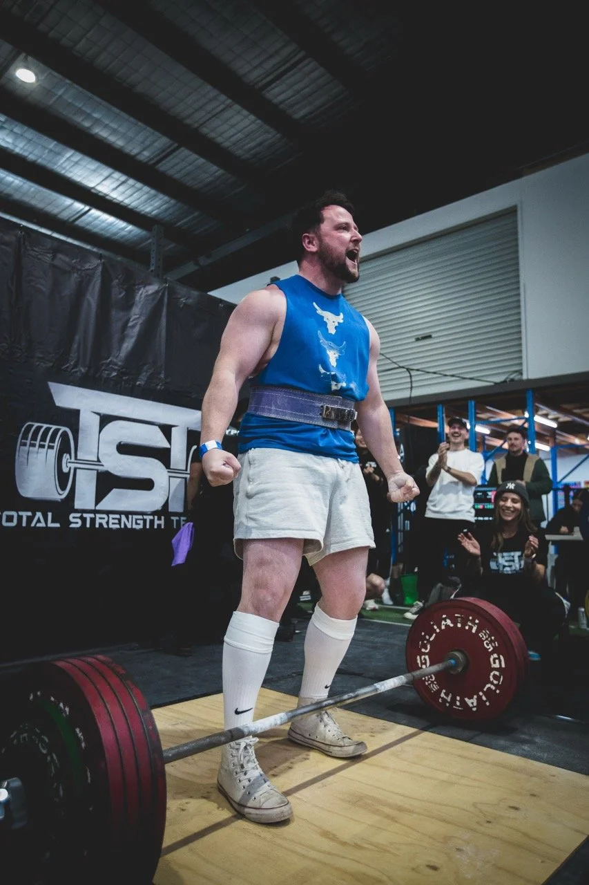 A man with a beard and tattoos lifting a barbell during a powerlifting event at a gym with spectators in the background.