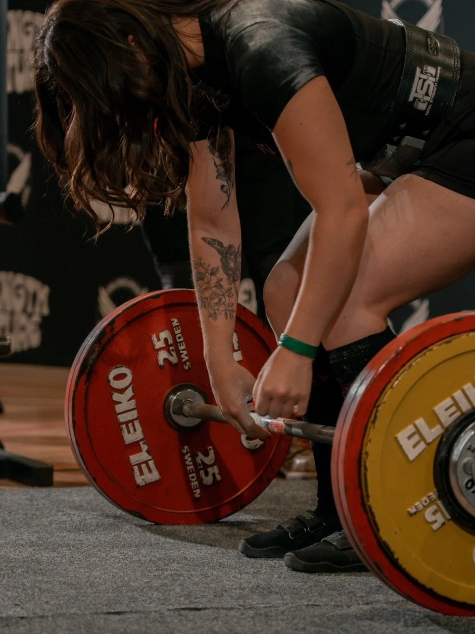 A woman with tattoos lifting a barbell with red and yellow weight plates during a powerlifting competition.