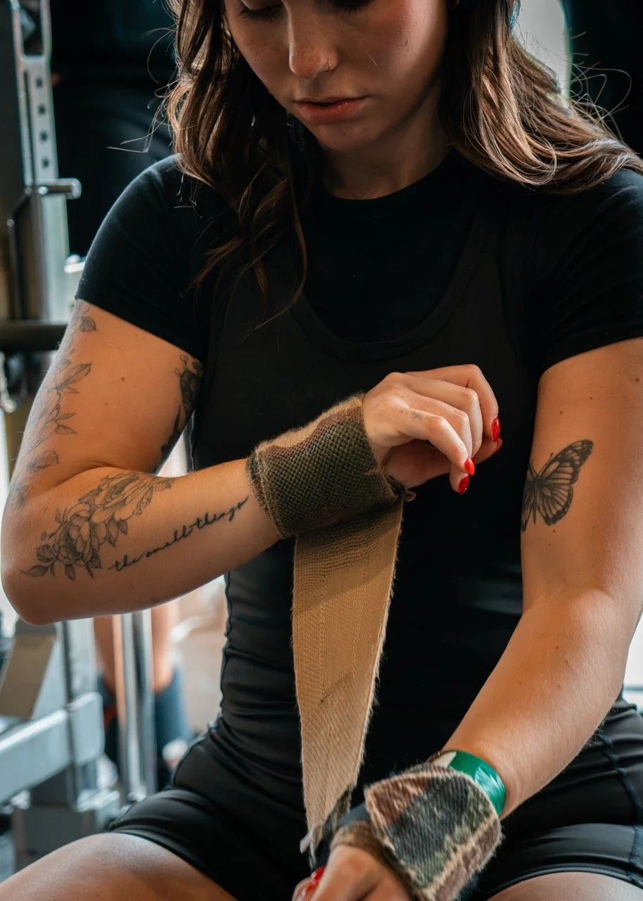 A young woman with tattoos on her arms is wrapping her wrist with a fabric band in a gym setting.