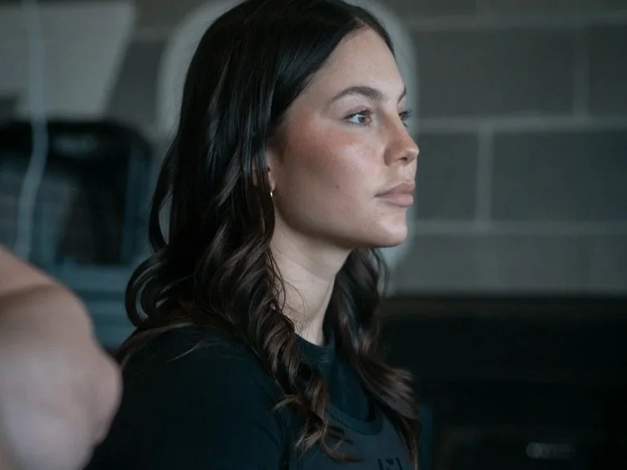 Two women in a gym, one whispering into the other’s ear, both in athletic clothing.