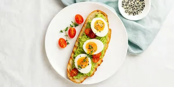 Open-faced avocado toast topped with sliced hard-boiled eggs and cherry tomatoes, served on a white plate.
