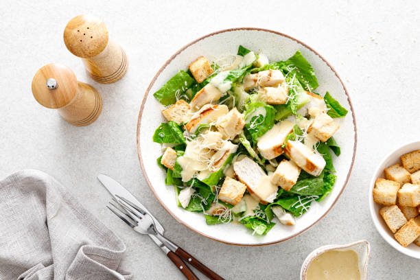 Caesar salad with romaine lettuce, croutons, and parmesan cheese in a white bowl, accompanied by salt and pepper shakers, a fork and knife, and a small bowl of salad dressing on a light gray surface.