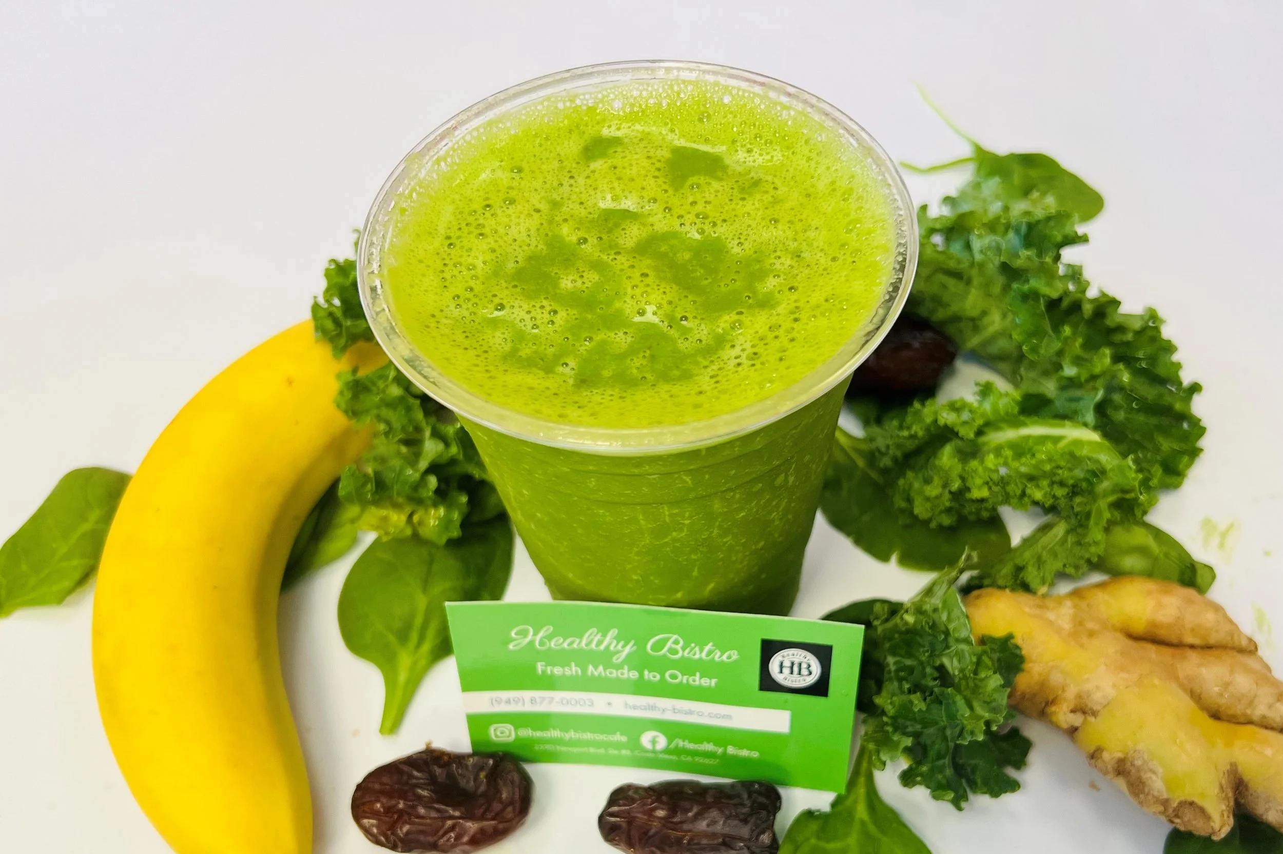 A green smoothie in a clear plastic cup topped with foam, surrounded by a banana, green leafy vegetables, ginger root, and dates, with a green business card reading 'Healthy Bistro'.