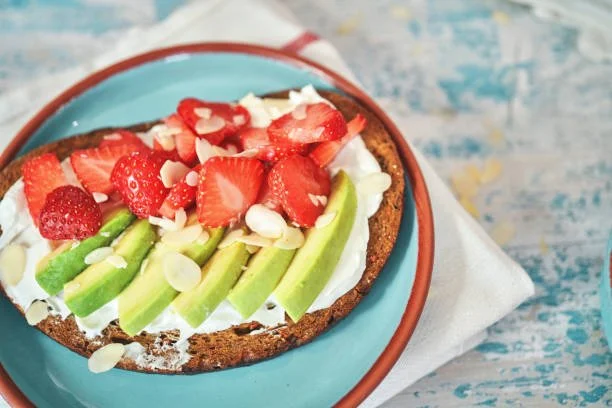 Slice of fruit tart topped with strawberries, avocado slices, and almond slices on a blue plate.