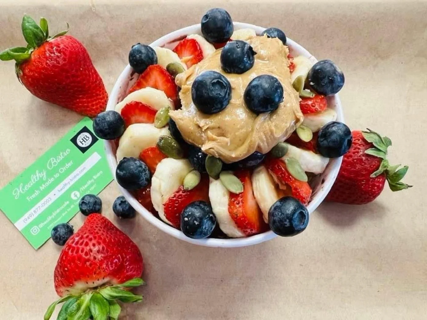 A bowl of mixed fresh strawberries, blueberries, banana slices, and pumpkin seeds topped with a dollop of peanut butter, surrounded by whole strawberries on a brown paper surface. A green business card with contact information is positioned next to t