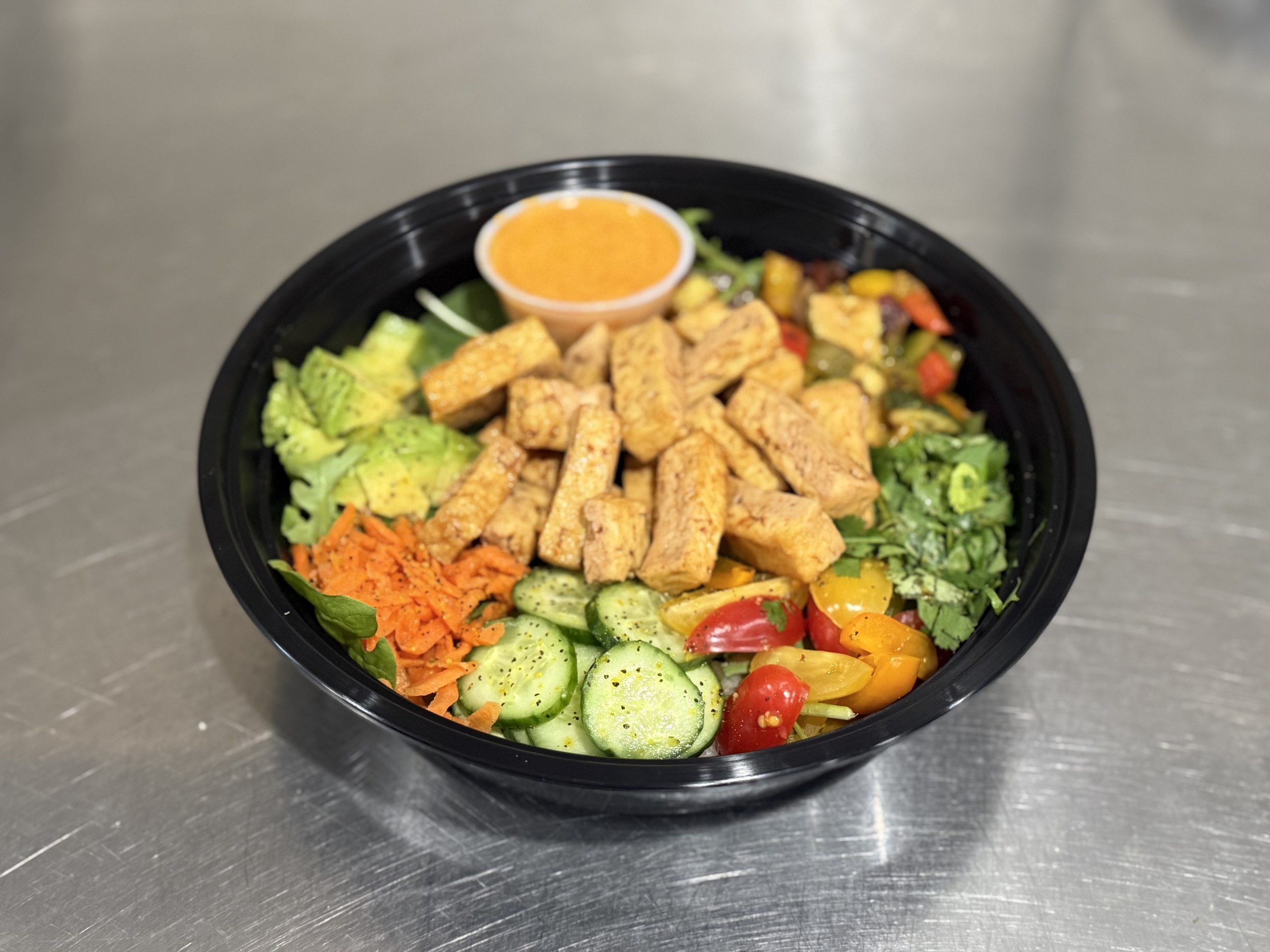 Salad with grilled chicken strips, cherry tomatoes, cucumber slices, shredded carrots, leafy greens, and a small container of orange dressing in a black bowl.