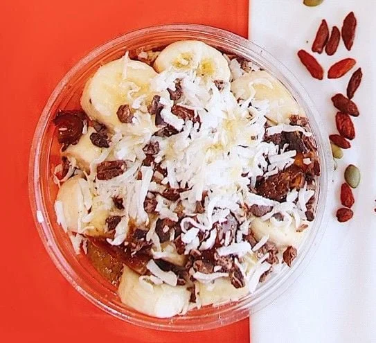 A clear plastic bowl of overnight oats with sliced bananas, shredded coconut, chopped nuts, and dried fruit, placed on a red and white background with some dried goji berries and pumpkin seeds beside it.