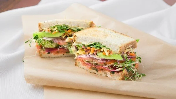 Close-up of a sandwich with bread, lettuce, tomato, avocado, sprouts, and deli meat on parchment paper.