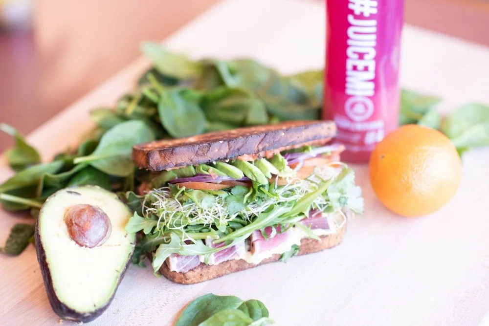 Open sandwich with vegetables and greens, avocado, orange, and a can of juice on a light-colored surface