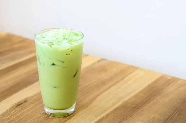A glass of iced matcha latte with ice cubes on a wooden surface.