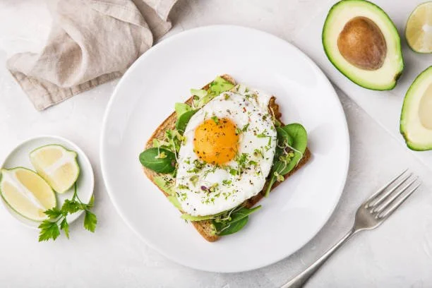 Open-faced sandwich with a fried egg on top, served on toast with greens, next to halved avocado, lemon wedges, and a sprig of parsley on a white plate.