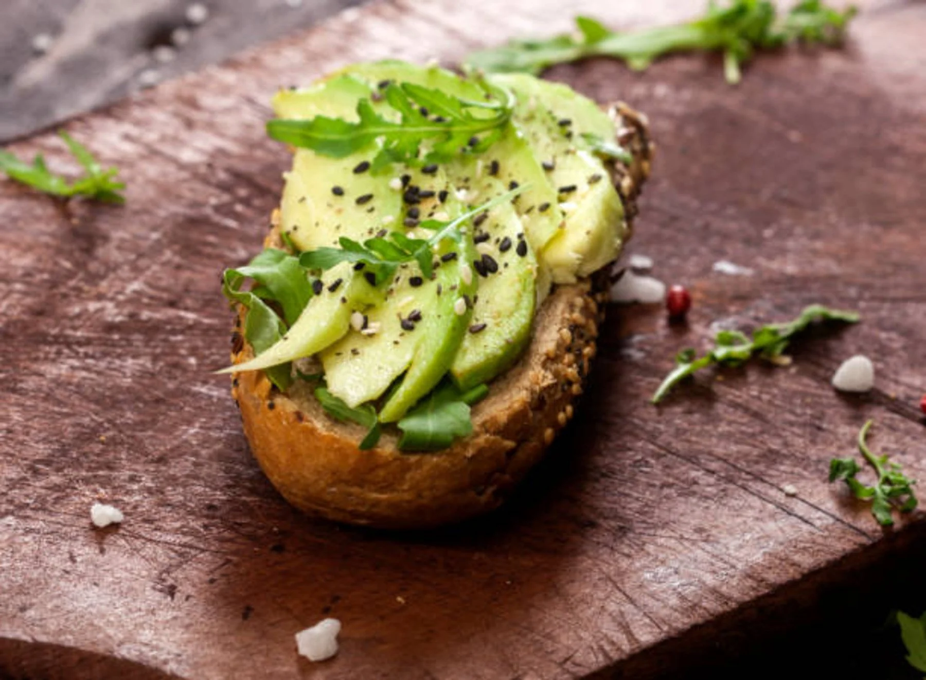 Open-faced avocado toast with sliced avocado, microgreens, black sesame seeds, and seasoning on a rustic wooden board.