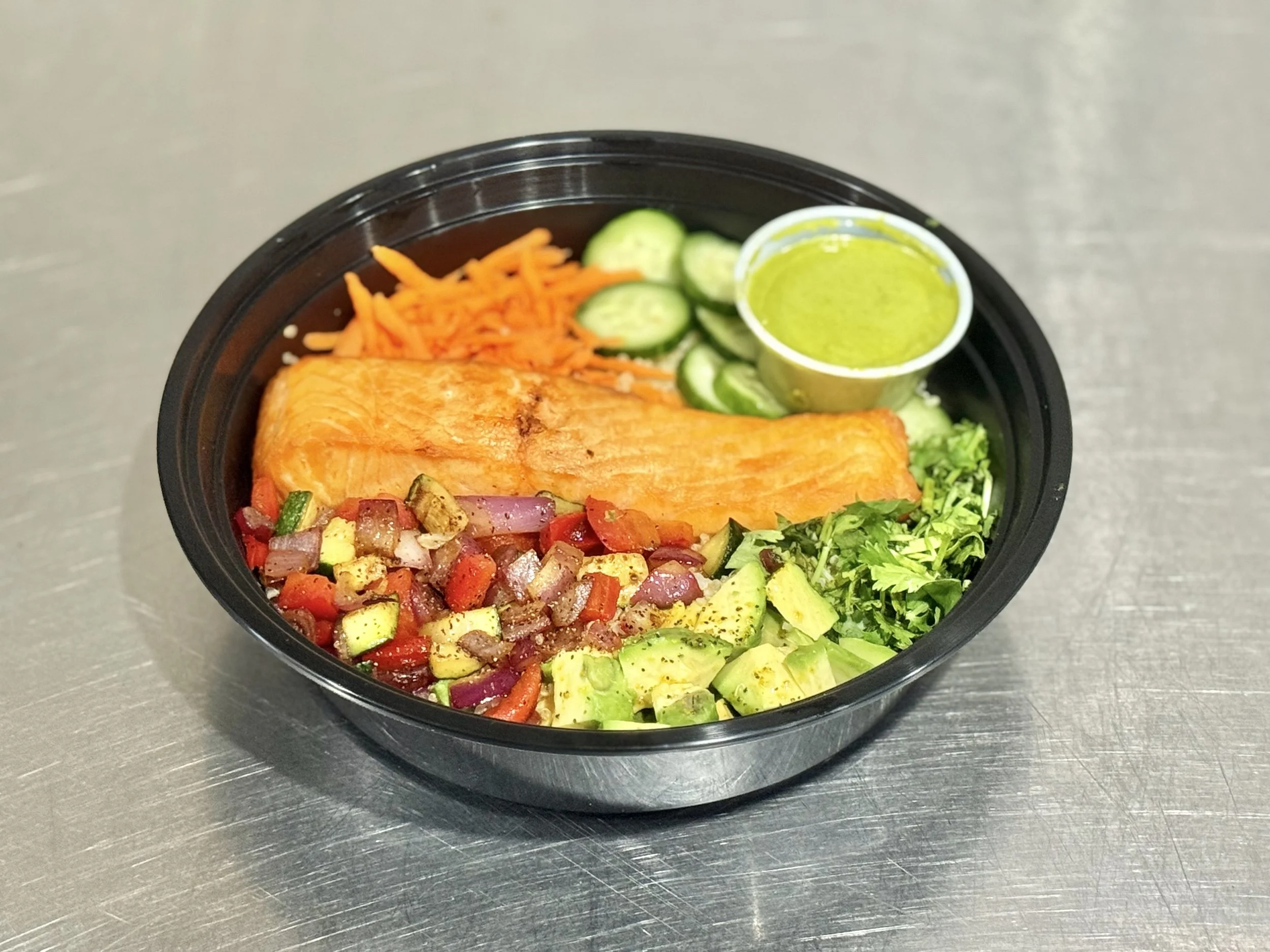 Salmon bowl with diced vegetables, shredded carrots, sliced cucumbers, chopped cilantro, and a side of green sauce in a small container.