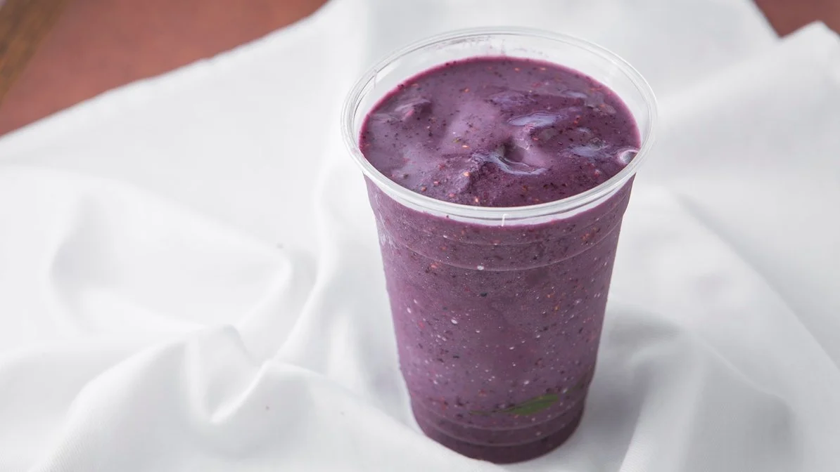 A clear plastic cup filled with a purple berry smoothie on a white cloth background.