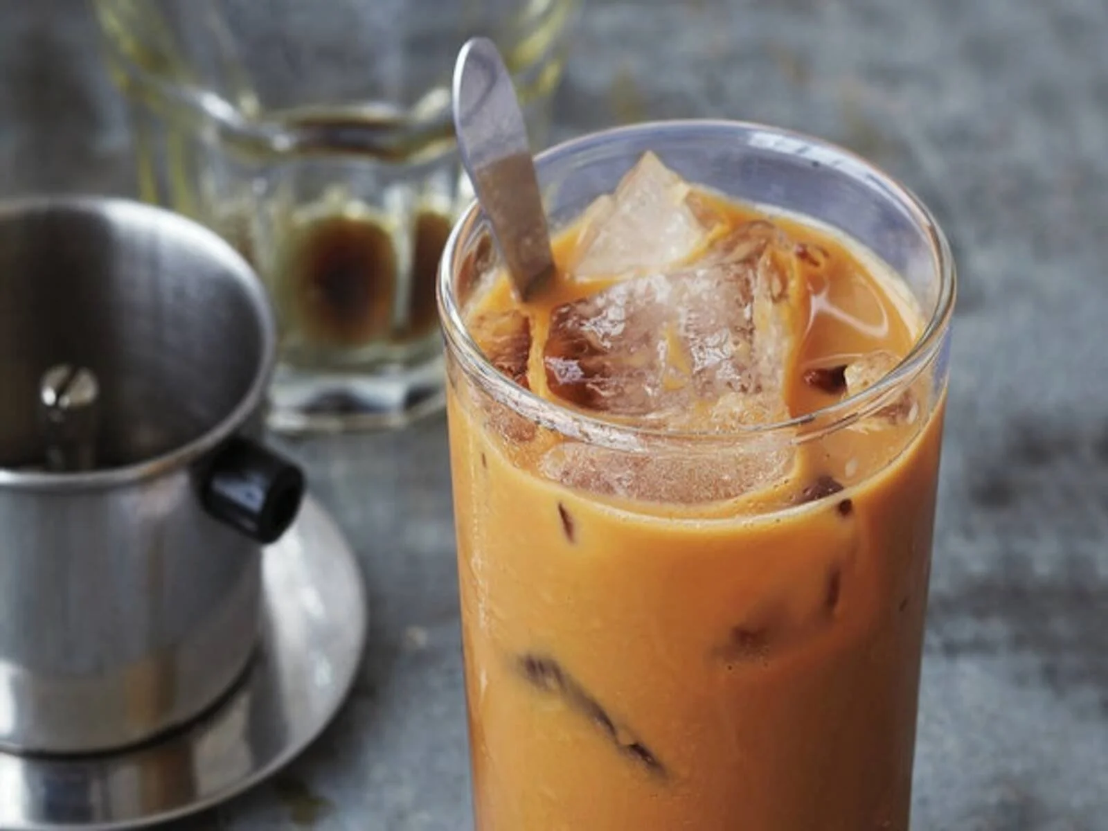 A tall glass of iced Thai iced tea with ice cubes, in a clear glass, on a wooden surface with a metal ice crusher and a jar of condensed milk in the background.