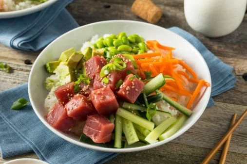 A bowl of poke with raw tuna, sliced cucumbers, shredded carrots, green peas, and chopped green onions on rice, served on a wooden table.