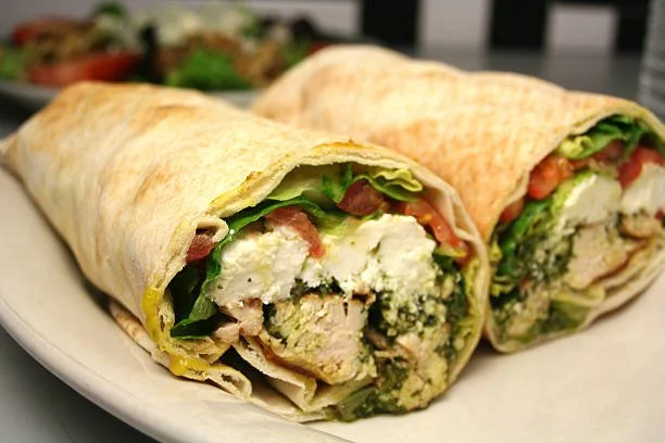 Close-up of two chicken Caesar wraps sliced in half, revealing lettuce, grilled chicken, and Caesar dressing inside a flour tortilla, on a white plate with a blurred salad in the background.