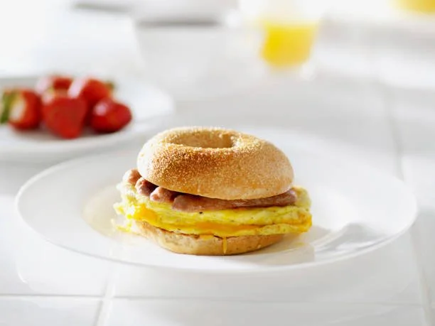 Bagel sandwich with scrambled eggs, ham, and cheese on a white plate with strawberries and a glass of orange juice in the background.