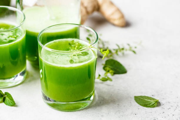 Three glasses of green juice on a white surface with fresh mint leaves and ginger in the background.
