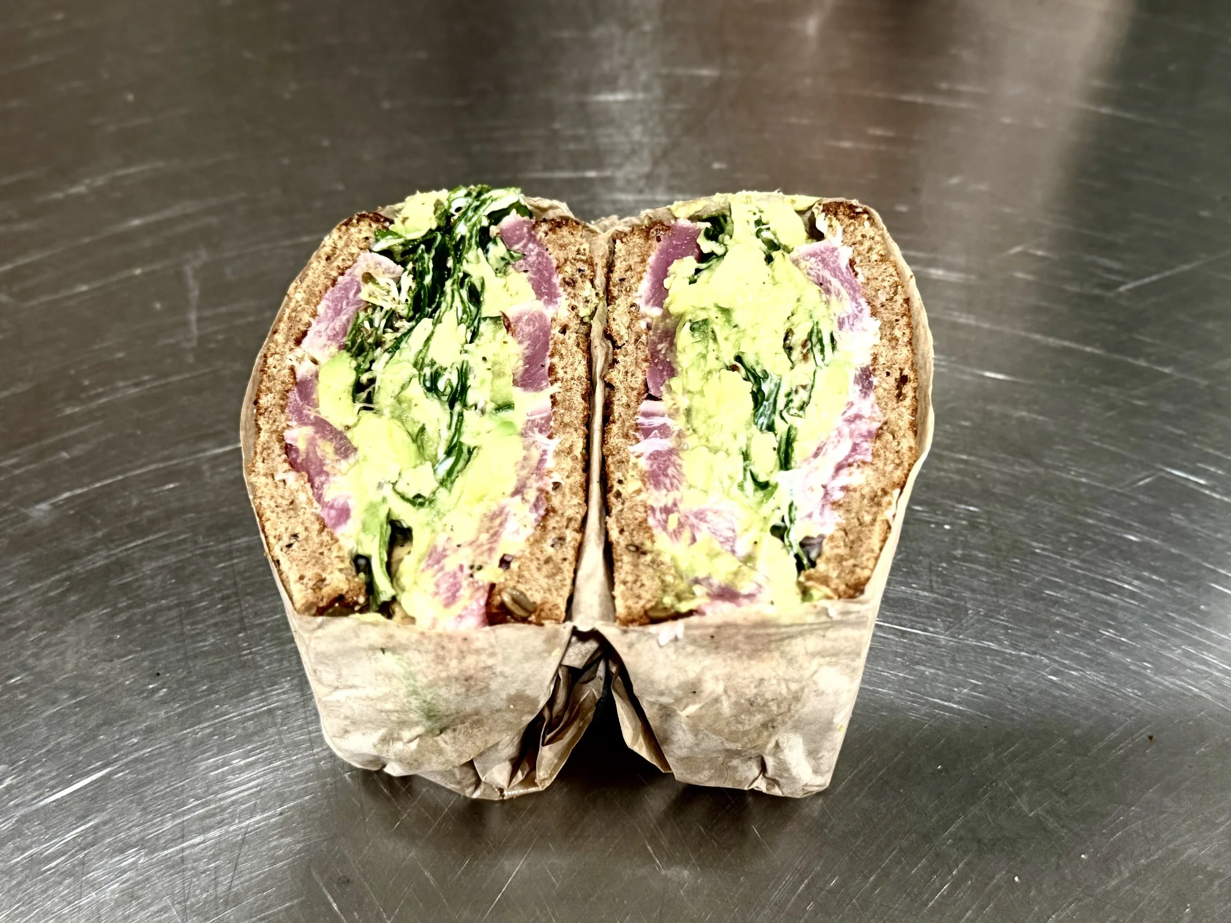 Half of a veggie sandwich with avocado, spinach, turkey, and mayonnaise in a wrapped paper on a metallic surface.