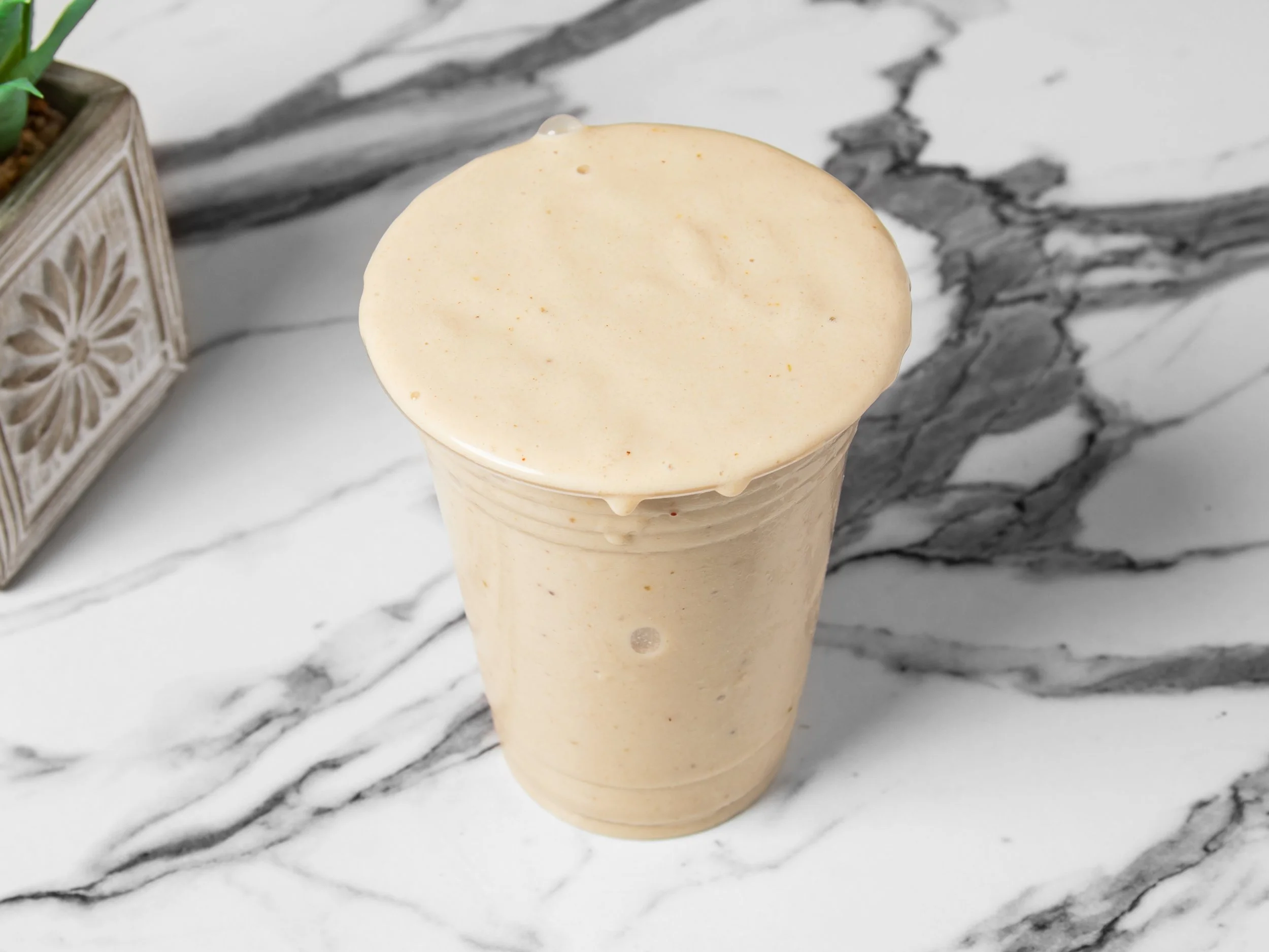 A creamy, frothy smoothie or milkshake in a clear plastic cup with a domed lid, placed on a marble surface.