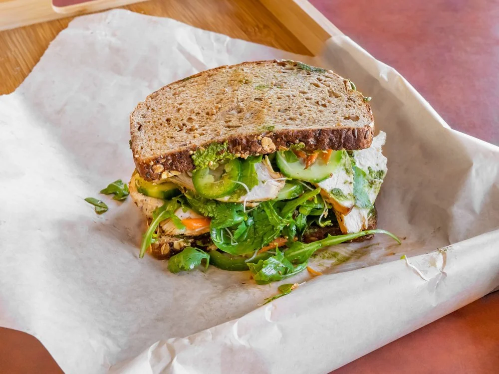 Vegetarian sandwich with whole grain bread, leafy greens, sliced cucumbers, and vegetables on white parchment paper.