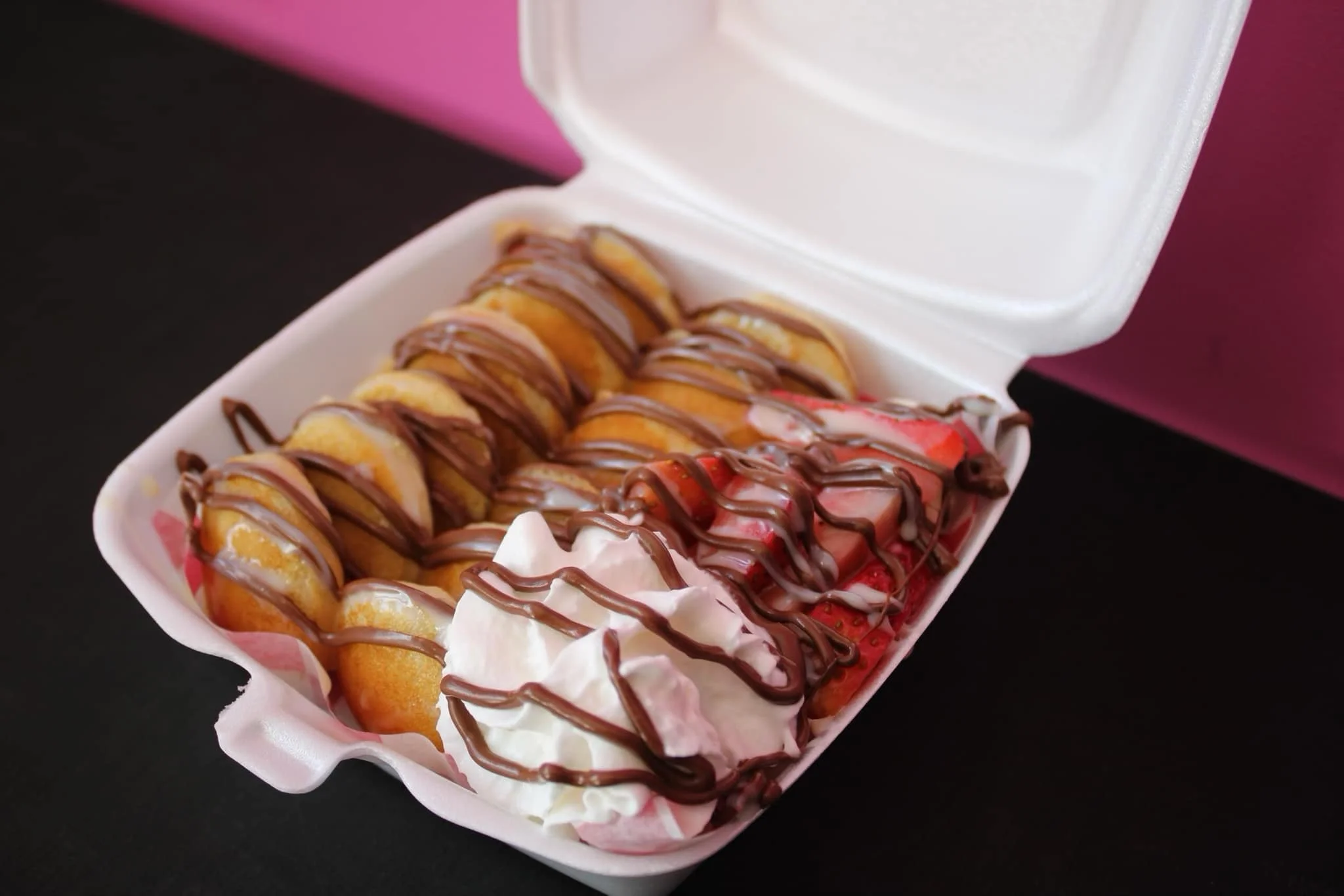 A takeout box containing sliced strawberries topped with strawberries or strawberry flavored ice cream, whipped cream, and drizzled with chocolate sauce.