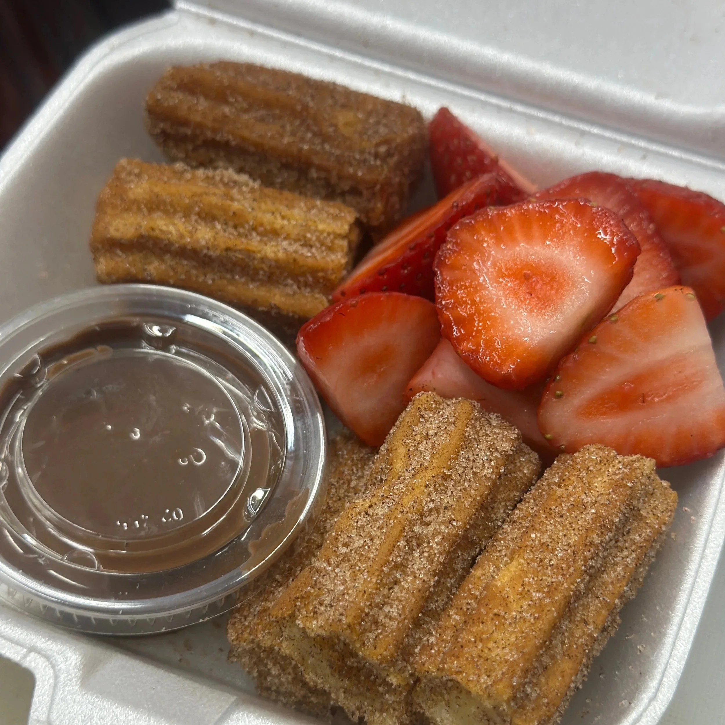 Churros with cinnamon sugar, sliced strawberries, and chocolate dipping sauce in a takeout container.
