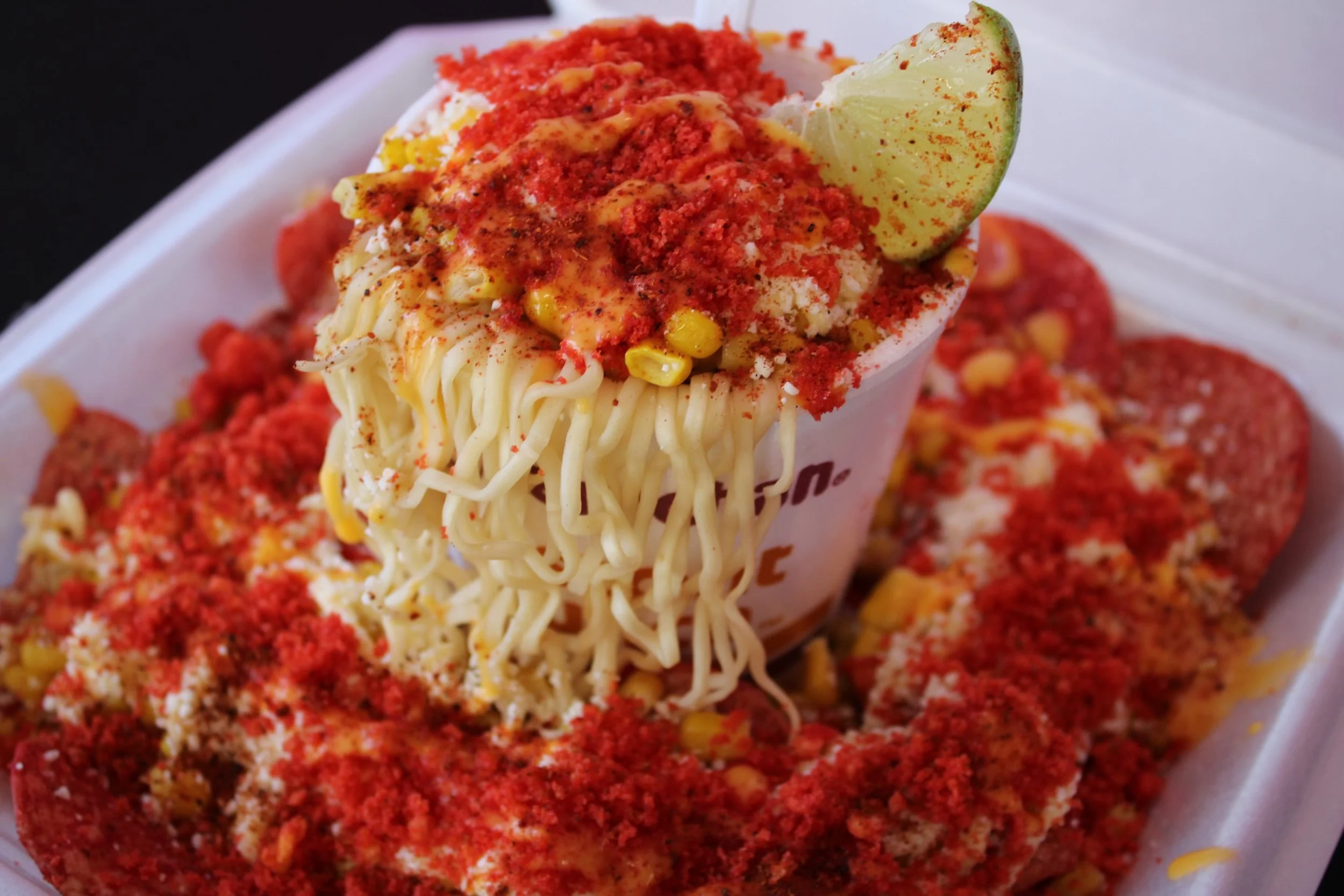 Close-up of a cup of instant ramen noodles topped with red seasoning and corn, with a lime wedge on the side, placed on a tray surrounded by fried chicken pieces.