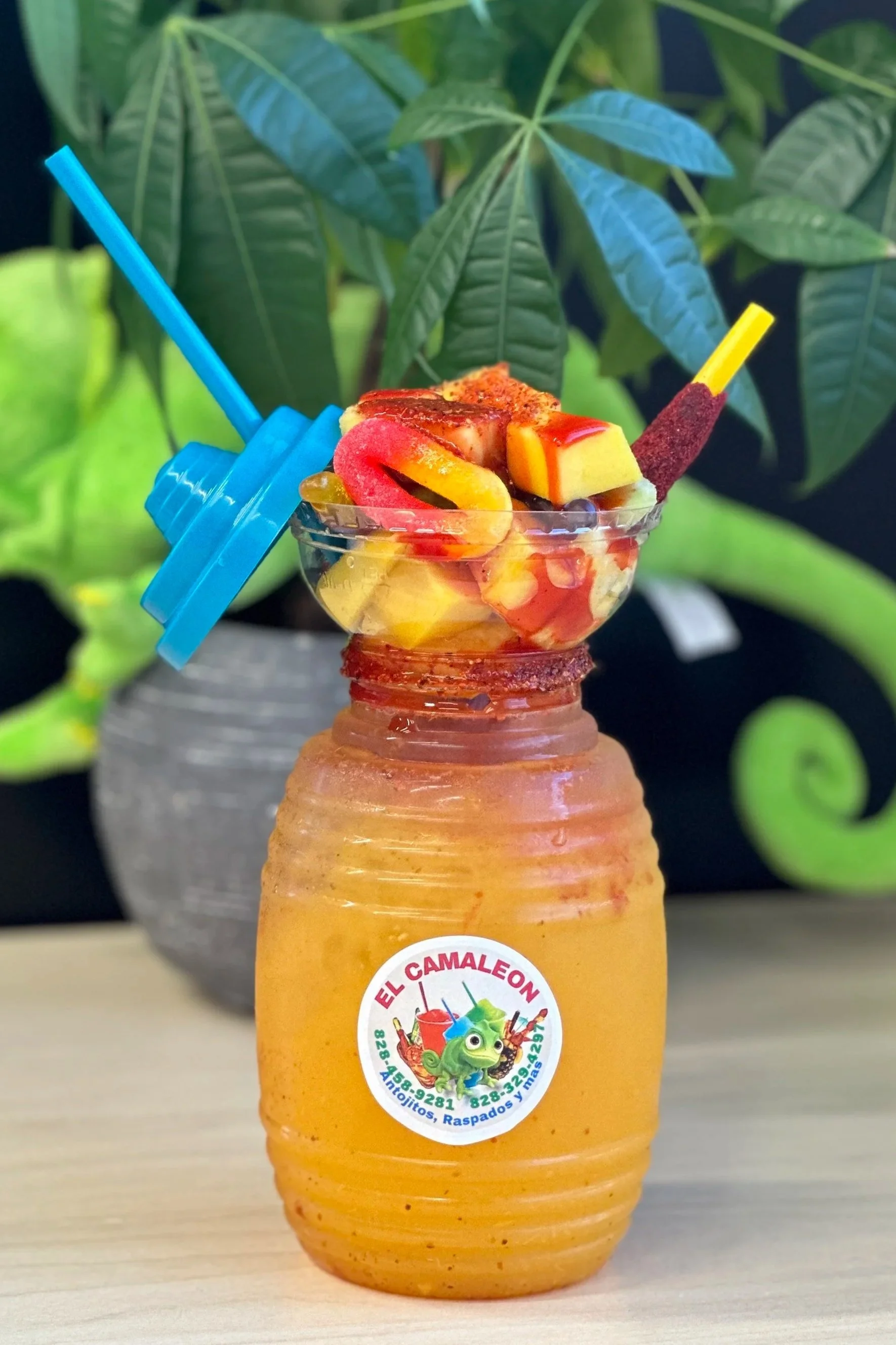 A jar of mango juice topped with sliced mango, lucas, and tamarind, with a bowl of fruit on top inside a plastic bowl, with a blue straw and a yellow and red candy stick, and a background of green plants.