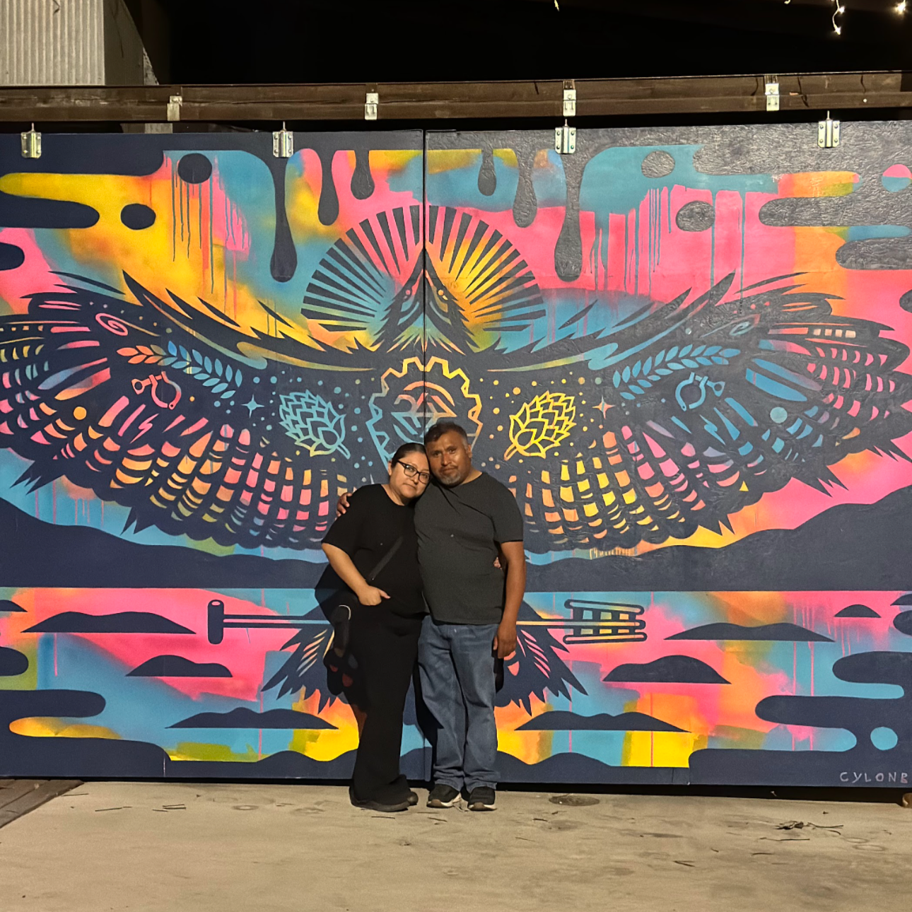 Two people standing in front of a colorful mural featuring a large butterfly with intricate wings, set against a vibrant background of pink, yellow, blue, and black with artistic drips and patterns.