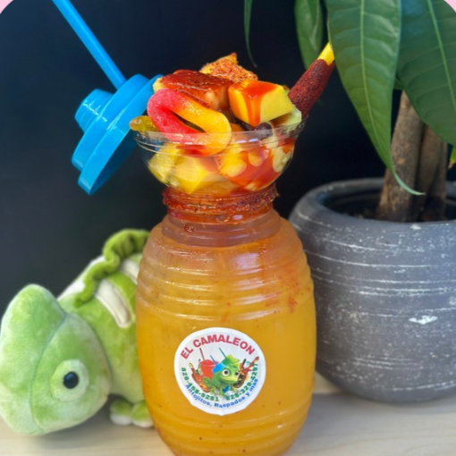 A barrel shaped cup  of Mexican Mango agua Fresca (fruit drink) with a cup of colorful fresh fruit and candy on top, garnished with a blue straw, placed next to a green plush chameleon toy and a potted plant.